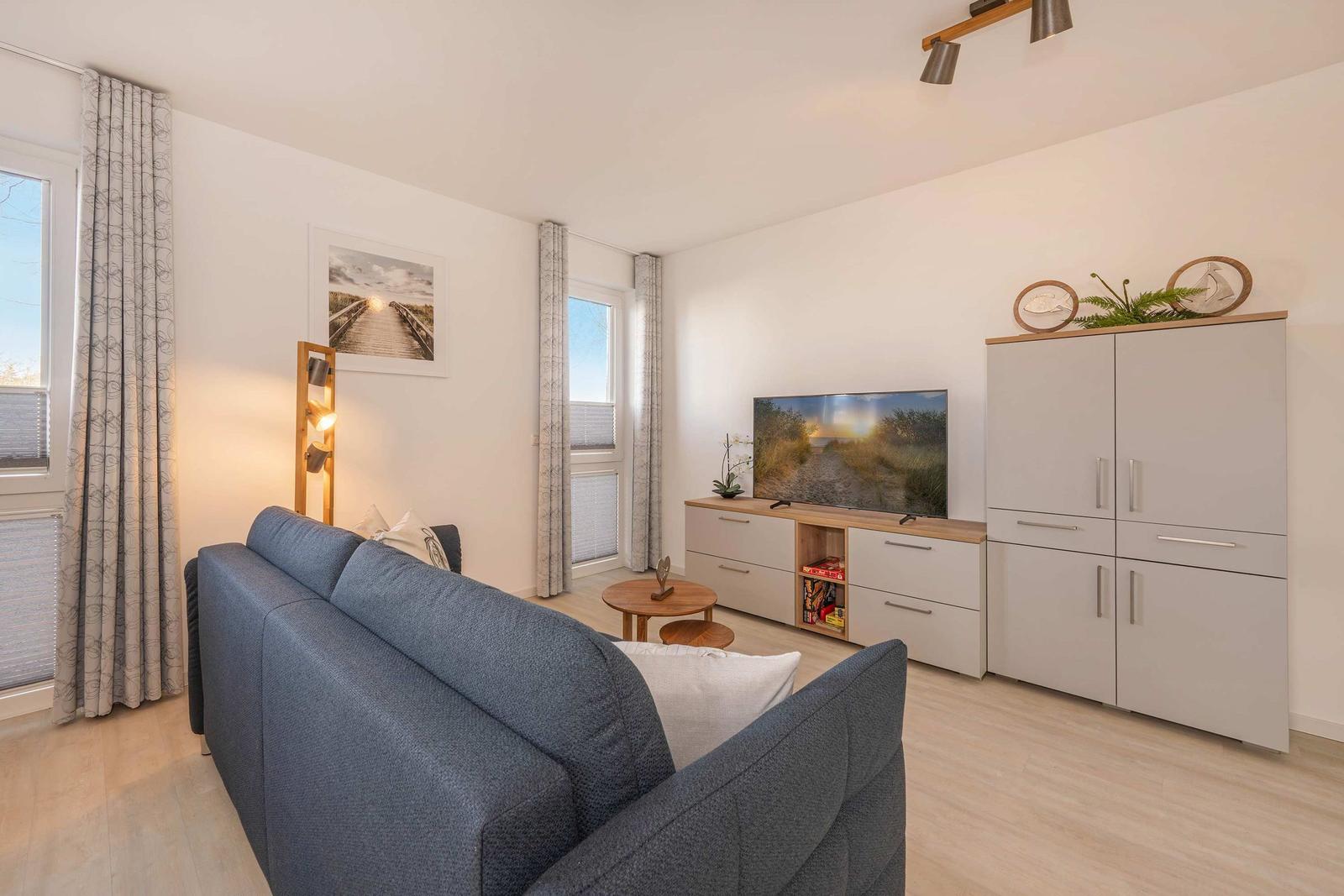 Ein Wohnzimmer mit Sofa, Fernseher und Schränken. Fenster mit Vorhängen.