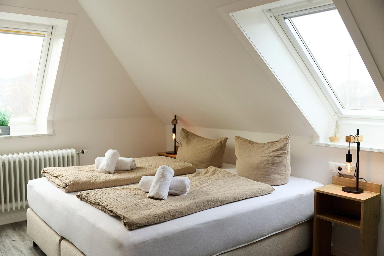Schlafzimmer mit Dachschrägen, Bett mit Kissen und Handtüchern, Fenster und Nachttisch.