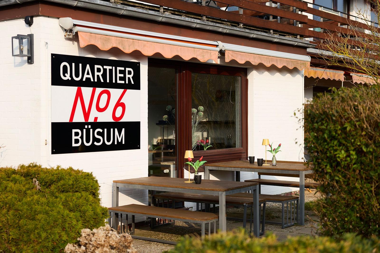 Außenbereich mit Tischen und Bänken vor einem Gebäude mit Schild QUARTIER N6 BÜSUM.