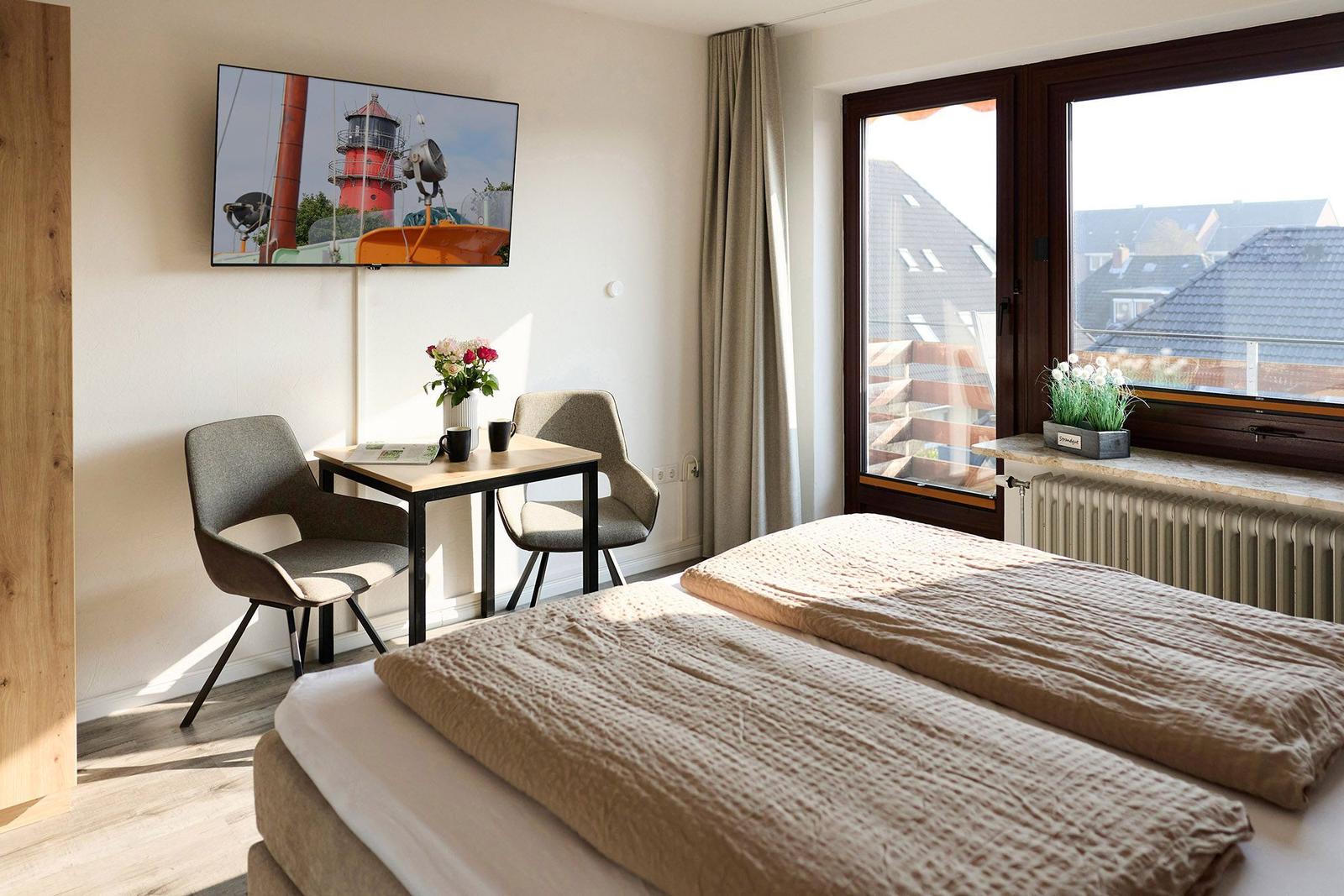 Zimmer mit Bett, Tisch, zwei Stühlen und TV. Fenster mit Blick auf Dächer.