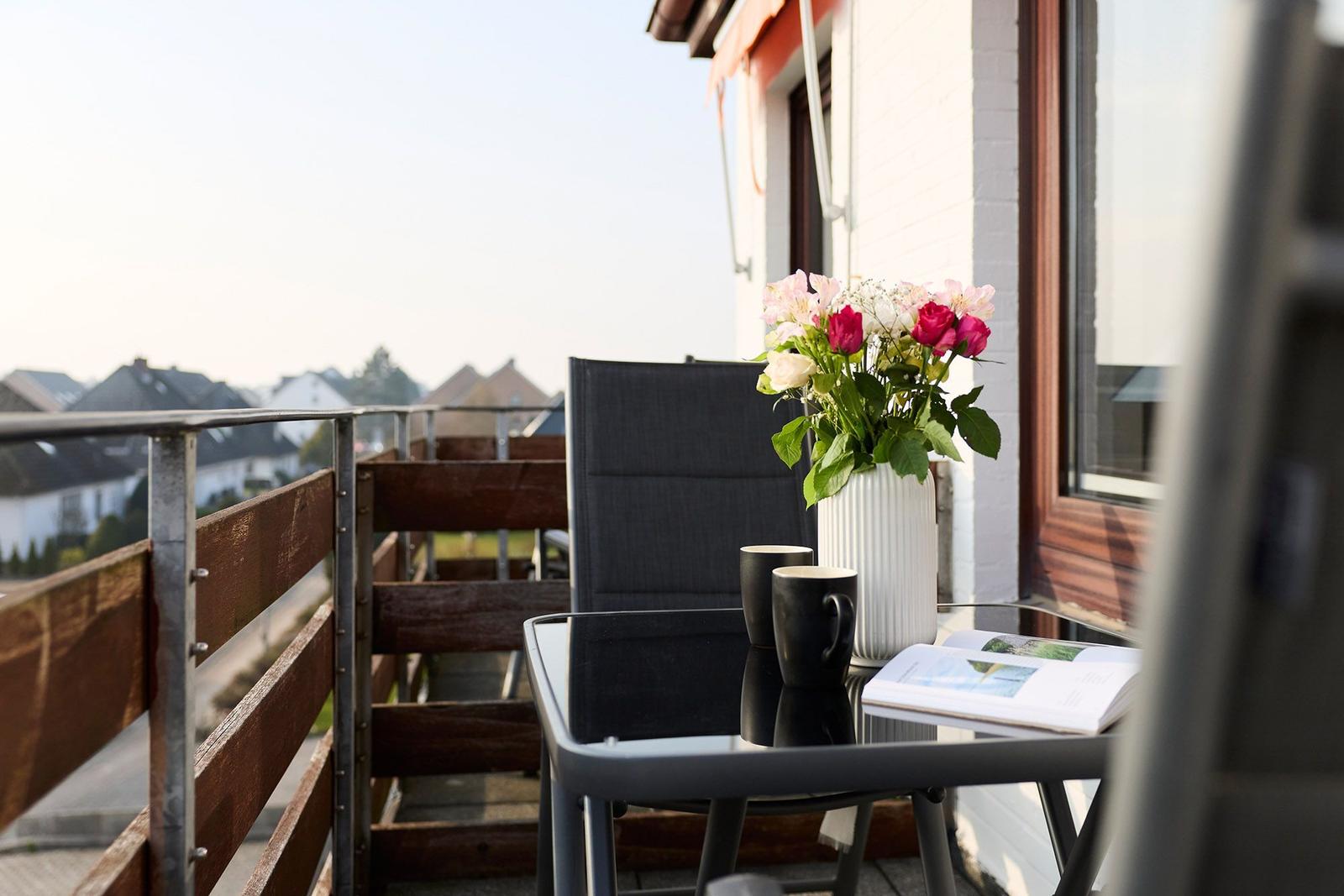 Balcony with table, chairs, and flower vase. View over rooftops and greenery.