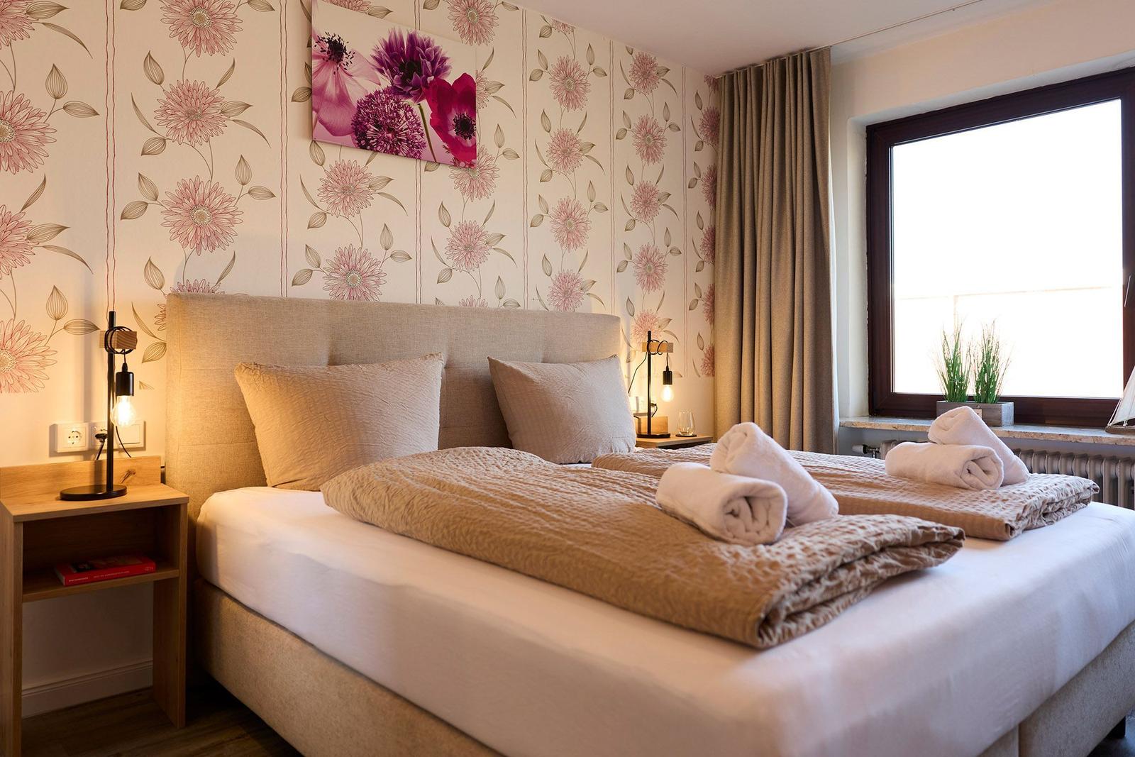 Doppelbett mit hellen Kissen, floraler Tapete und Fenster mit Blick.