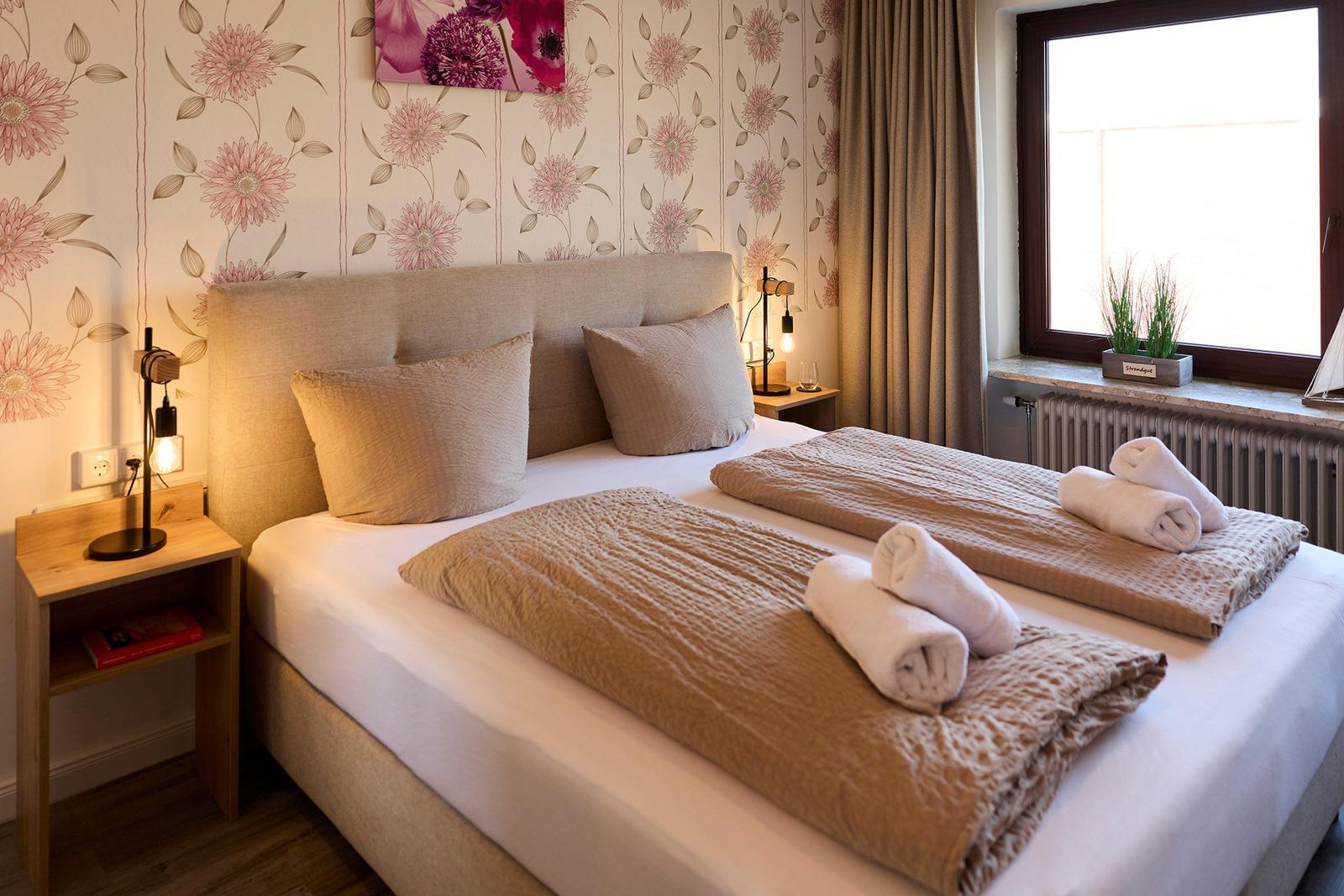 Doppelbett mit Kissen, Bettwäsche und Handtüchern. Blumendekor an der Wand und Fenster mit Vorhängen.