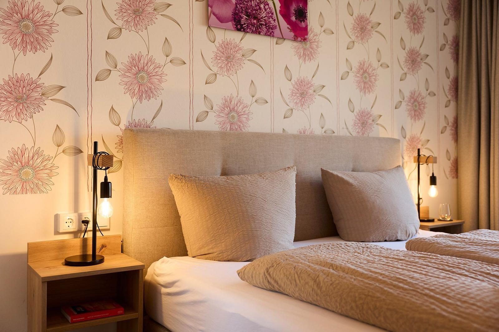 Doppelbett mit Kopfkissen, Nachttisch und Wandtapete mit Blumenmotiv.