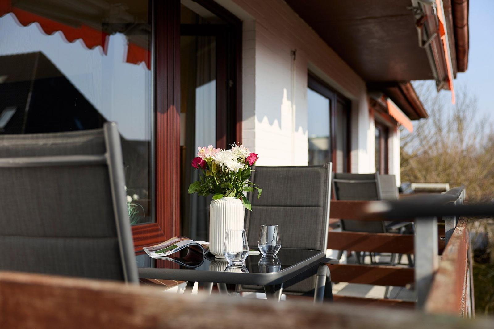 Terrasse mit Tisch, Stühlen und Blumenvase. Holzgeländer und Fenster sichtbar.