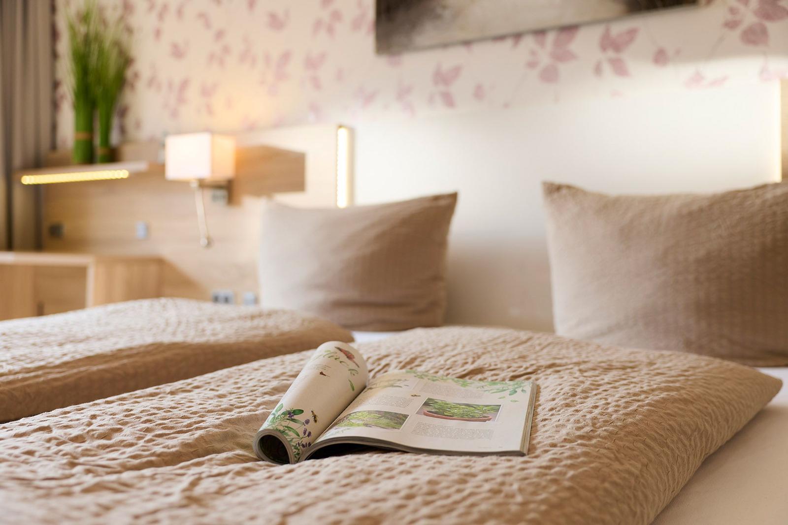 Ein Bett mit Kissen und einem Buch darauf. Wand mit Blumenmuster und Nachttischlampe.