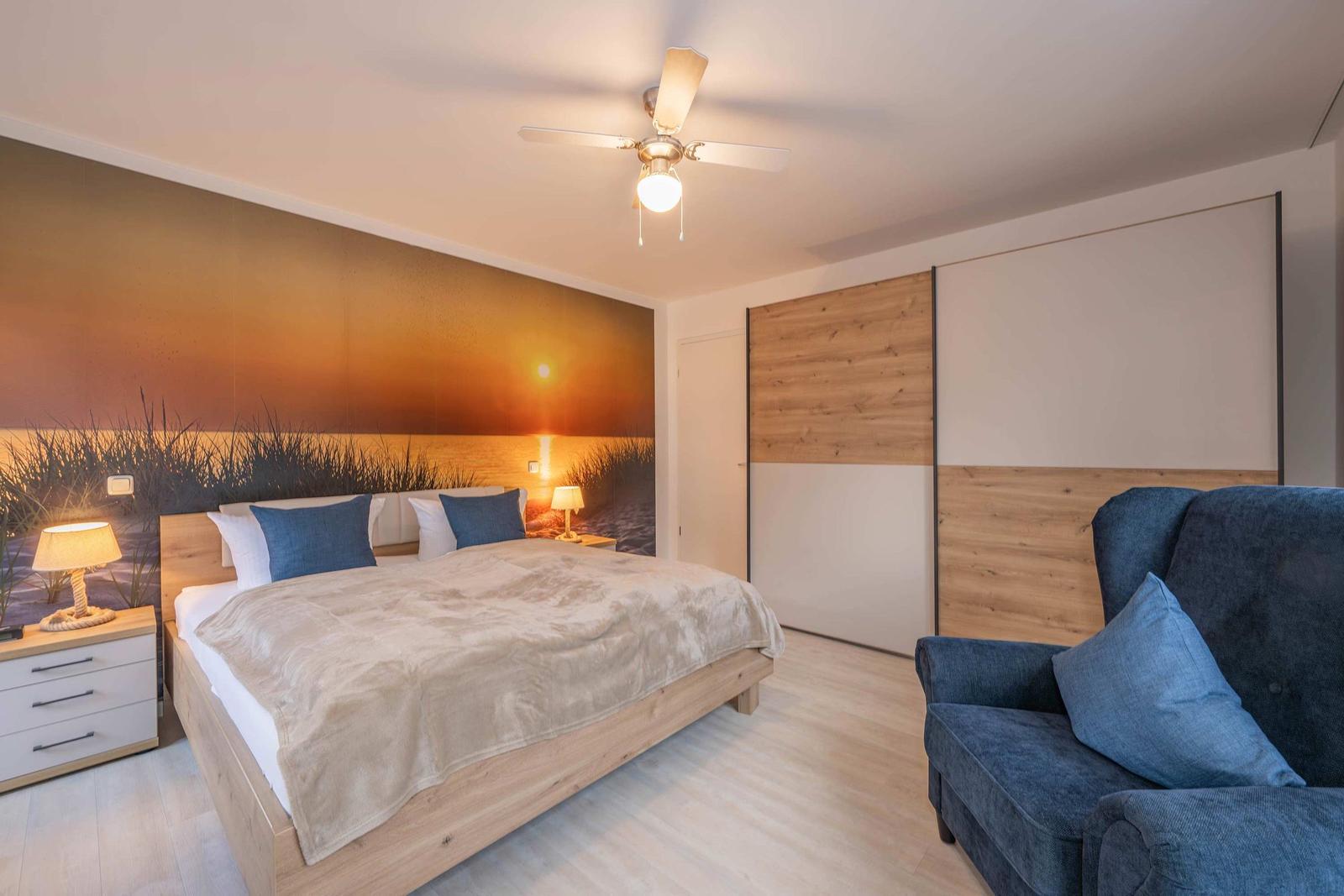 Schlafzimmer mit Bett, Sessel und Wandbild. Holzboden und Deckenventilator.