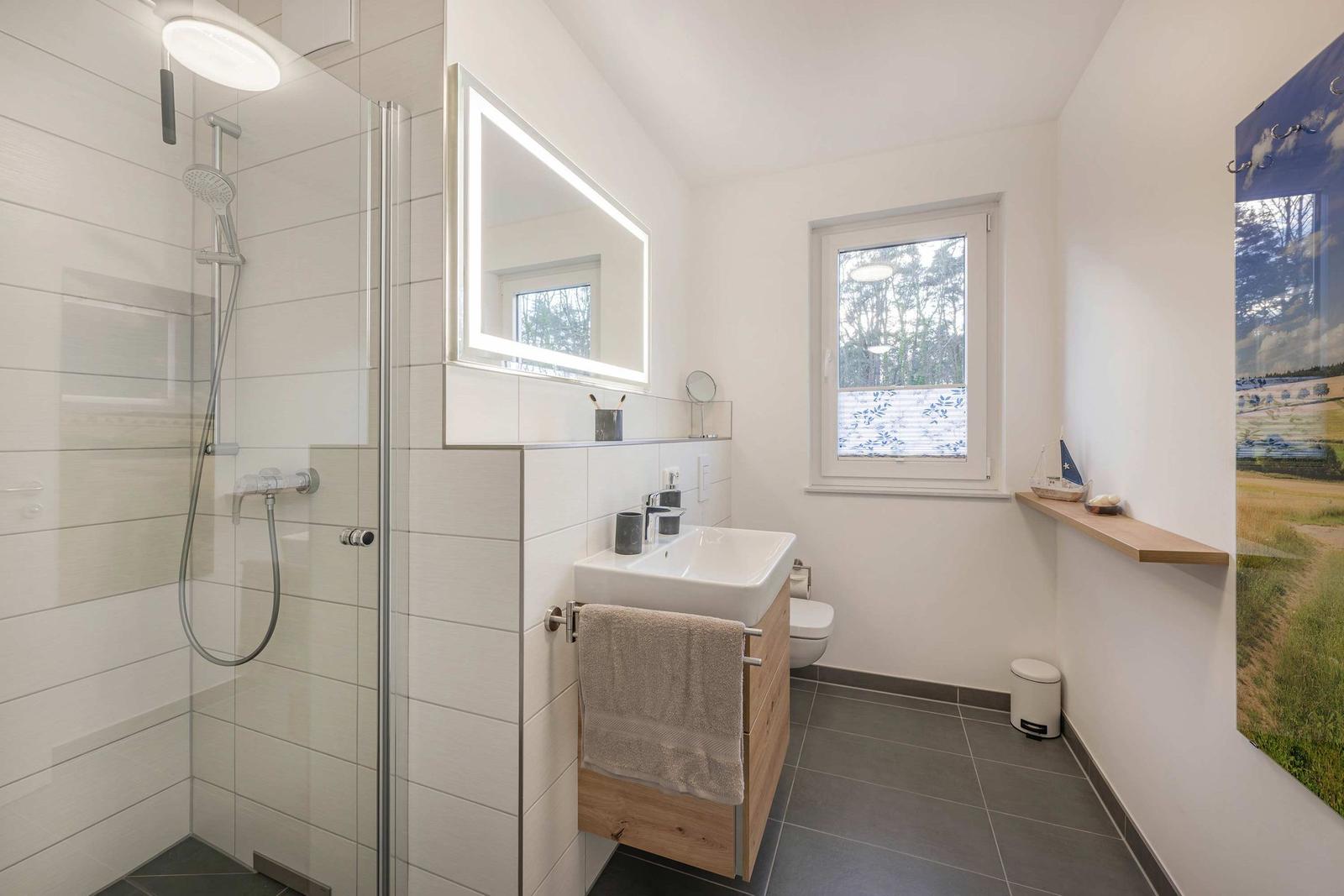 Badezimmer mit Dusche, Waschbecken und Toilette. Fenster mit Blick auf Wald.