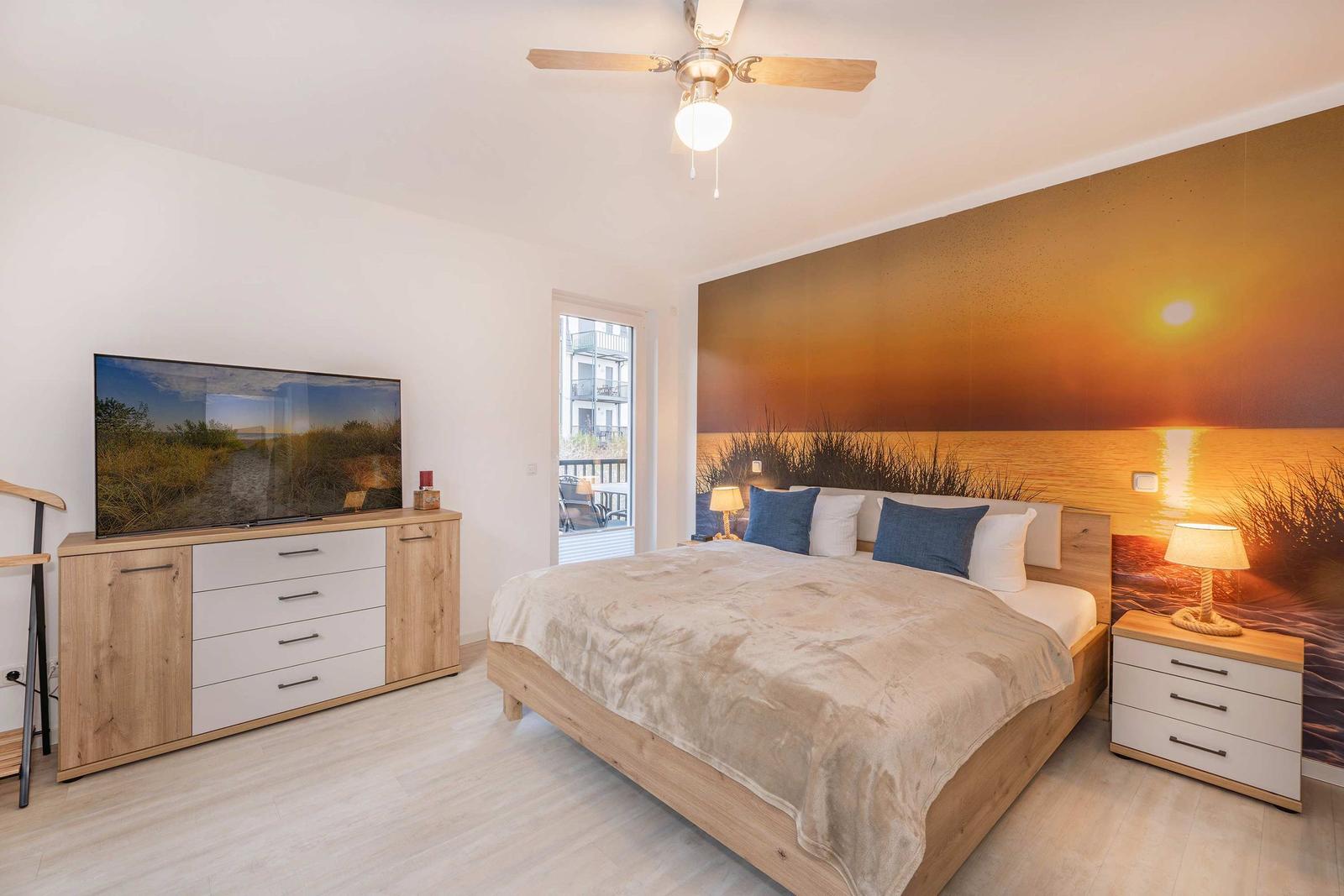 Schlafzimmer mit Bett, Nachttisch, TV-Schrank und Wandbild. Fenster mit Blick auf Balkon.