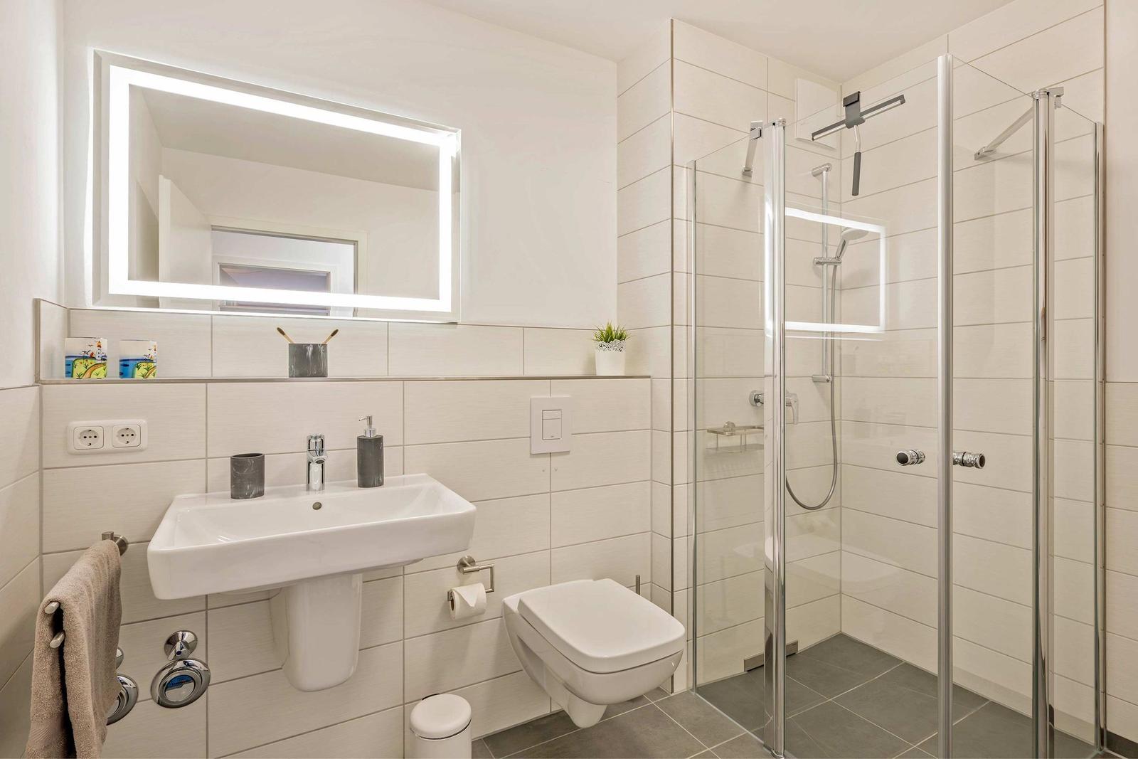 Badezimmer mit Dusche, Waschbecken und Toilette. Beleuchtetes Spiegel und graue Fliesen.