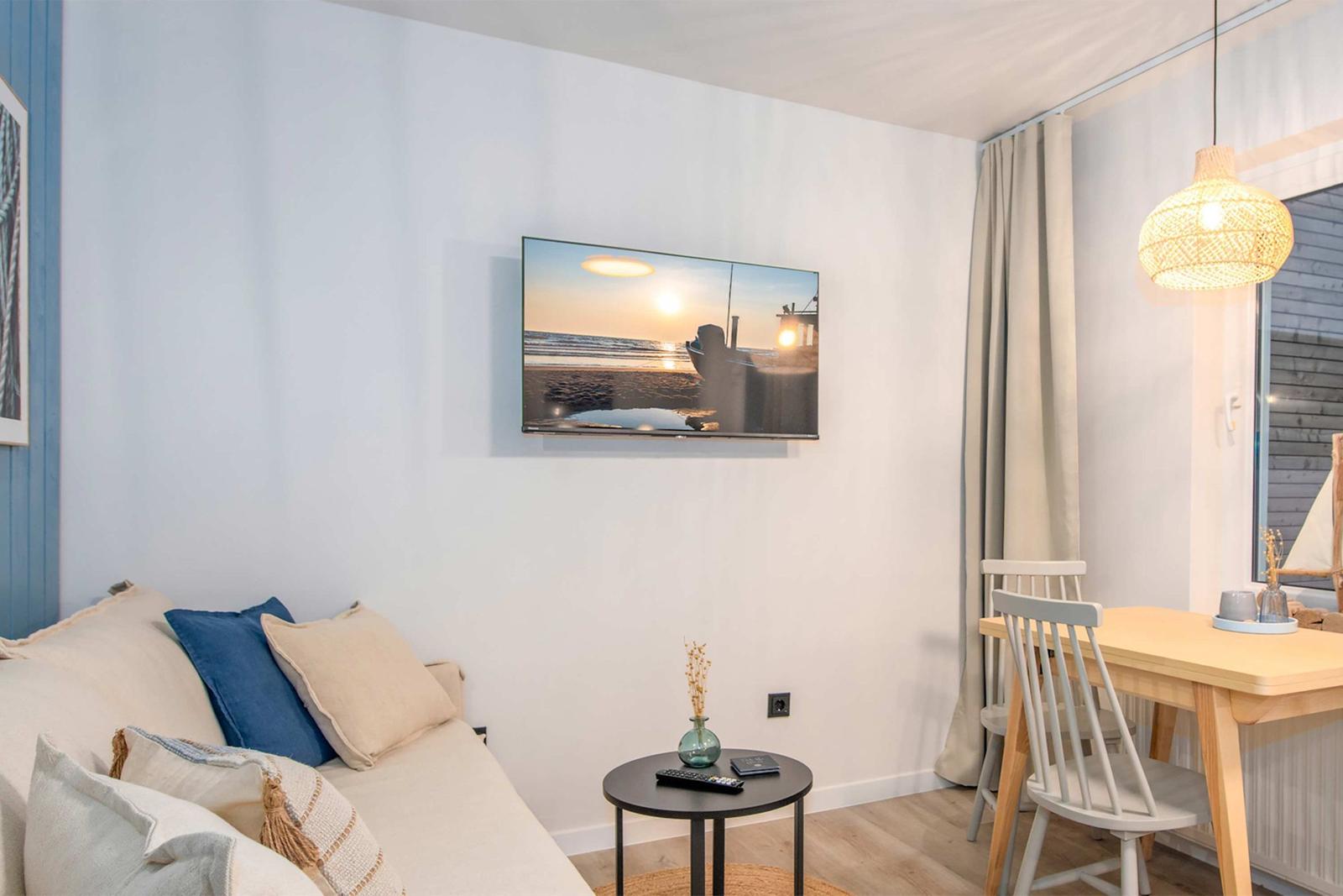 Wohnbereich mit Sofa, TV und Esstisch. Wandbild zeigt Sonnenuntergang.