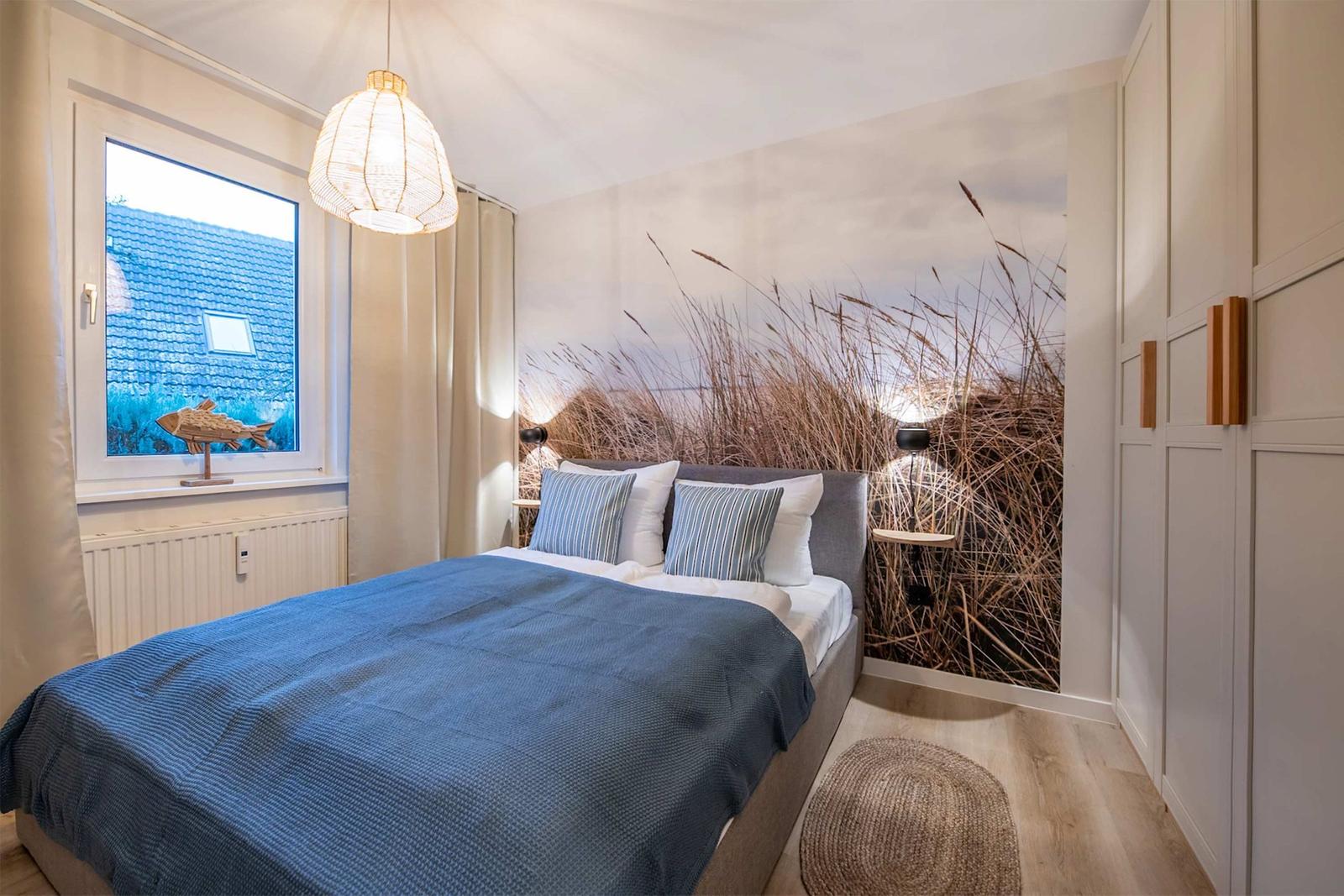 Schlafzimmer mit Bett, Wandbild von Schilf und Fensterblick auf Dach.
