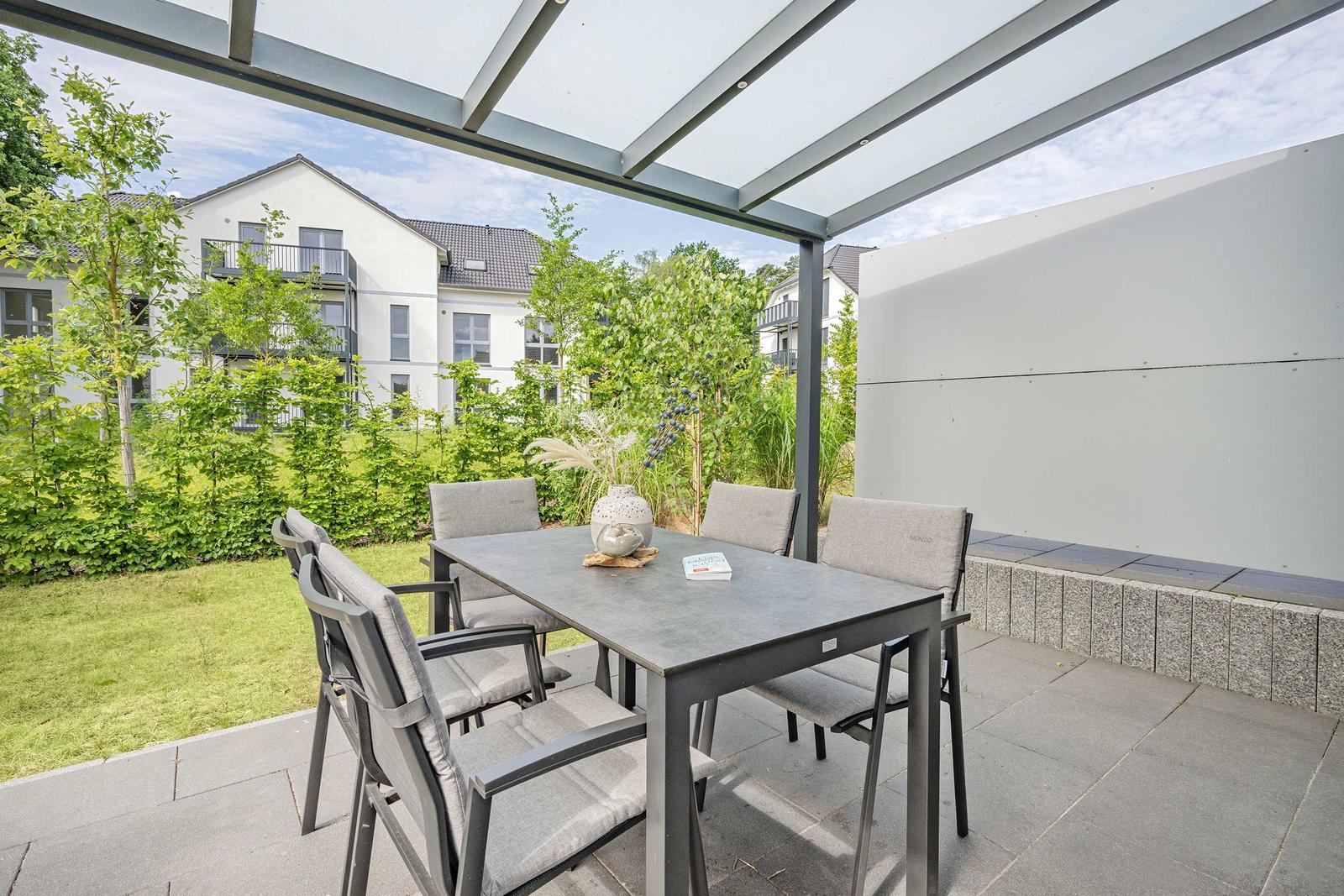 Terrasse mit Tisch und Stühlen unter Glasdach. Grünfläche und moderne Häuser im Hintergrund.