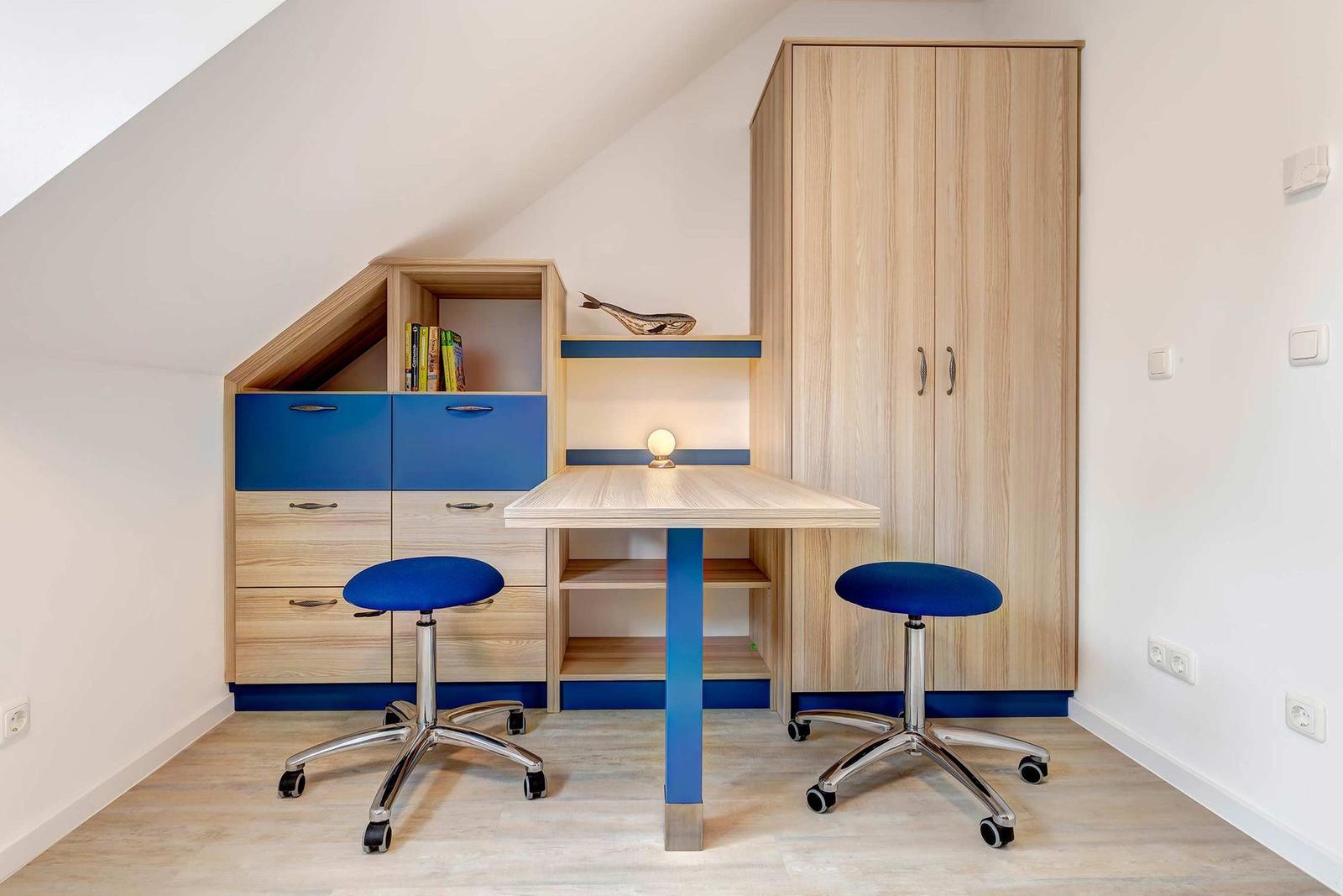 Ein Arbeitszimmer mit Schreibtisch, zwei blauen Stühlen und einem Holzschrank unter Dachschrägen.