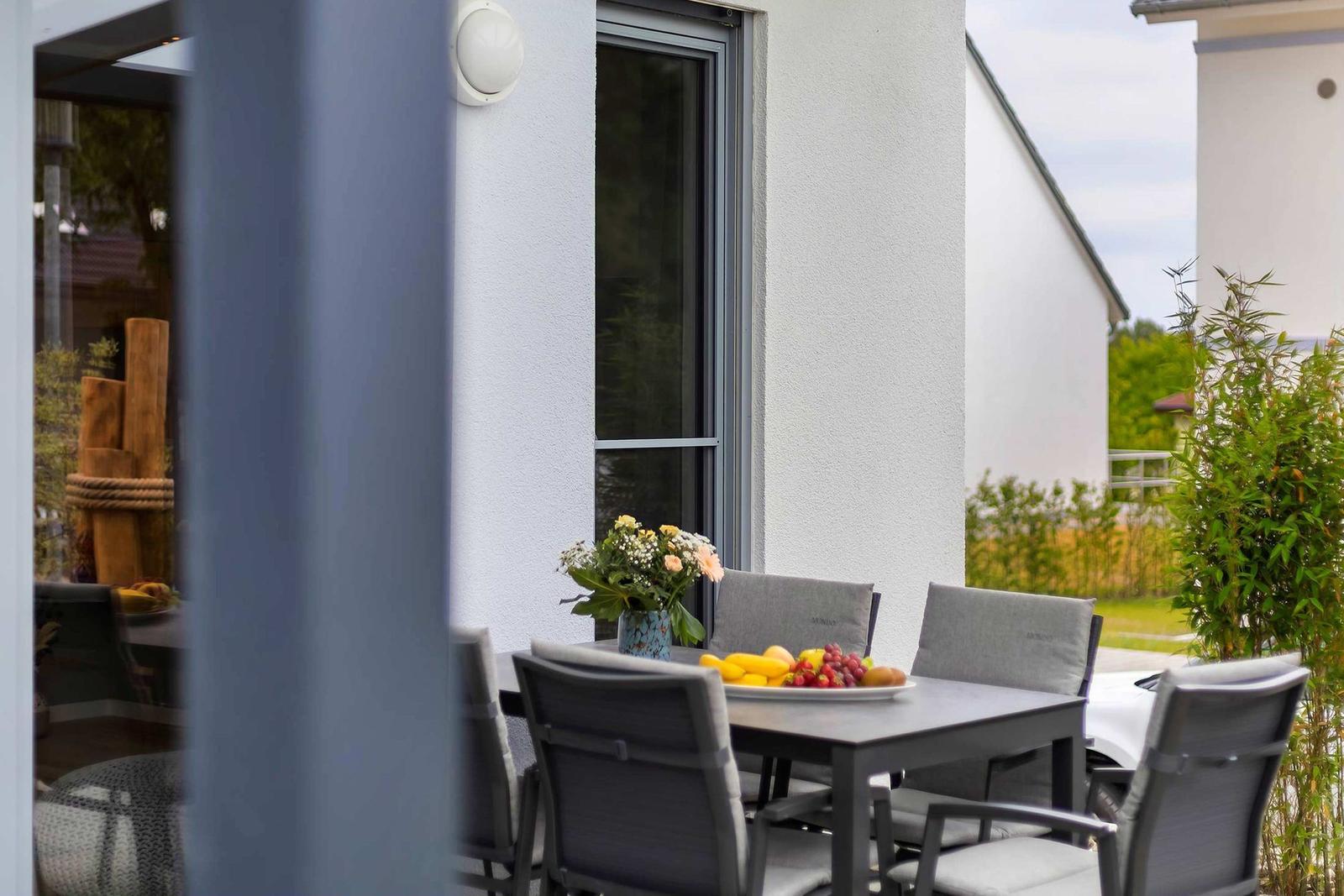 Terrasse mit Tisch, Stühlen und Obstschale. Hintergrund: Hauswand und Grünflächen.