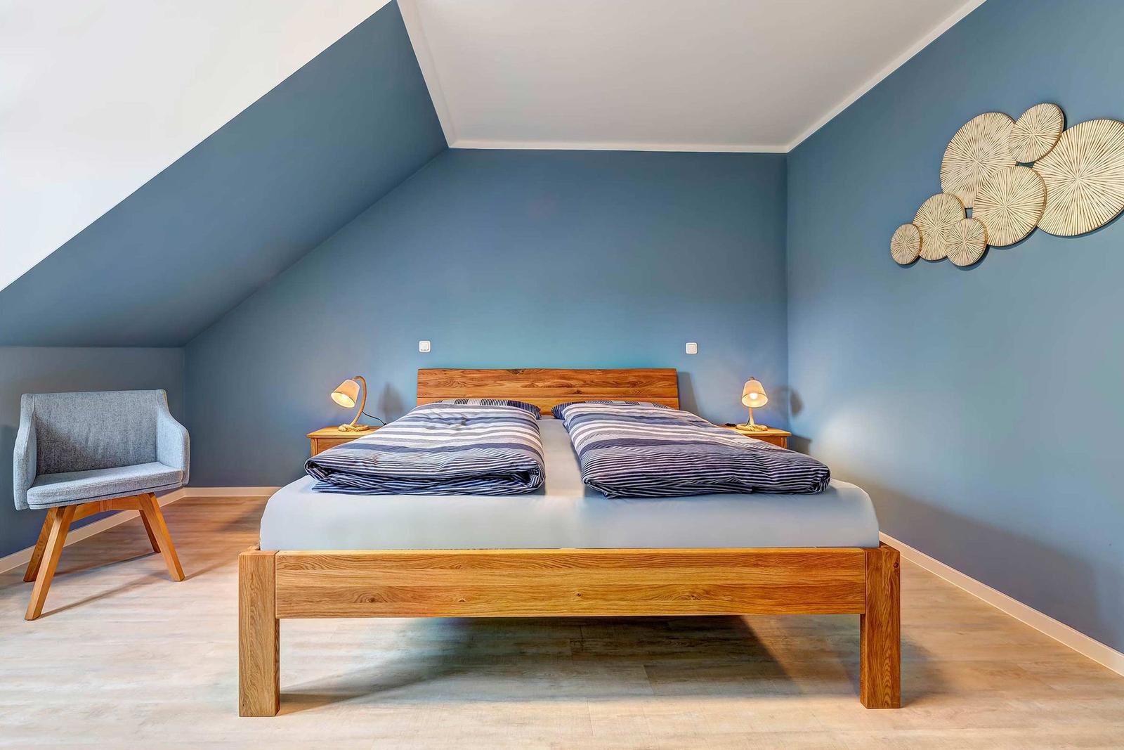 Doppelbett mit Holzrahmen, Nachttische und Sessel in einem Dachzimmer mit blauen Wänden.