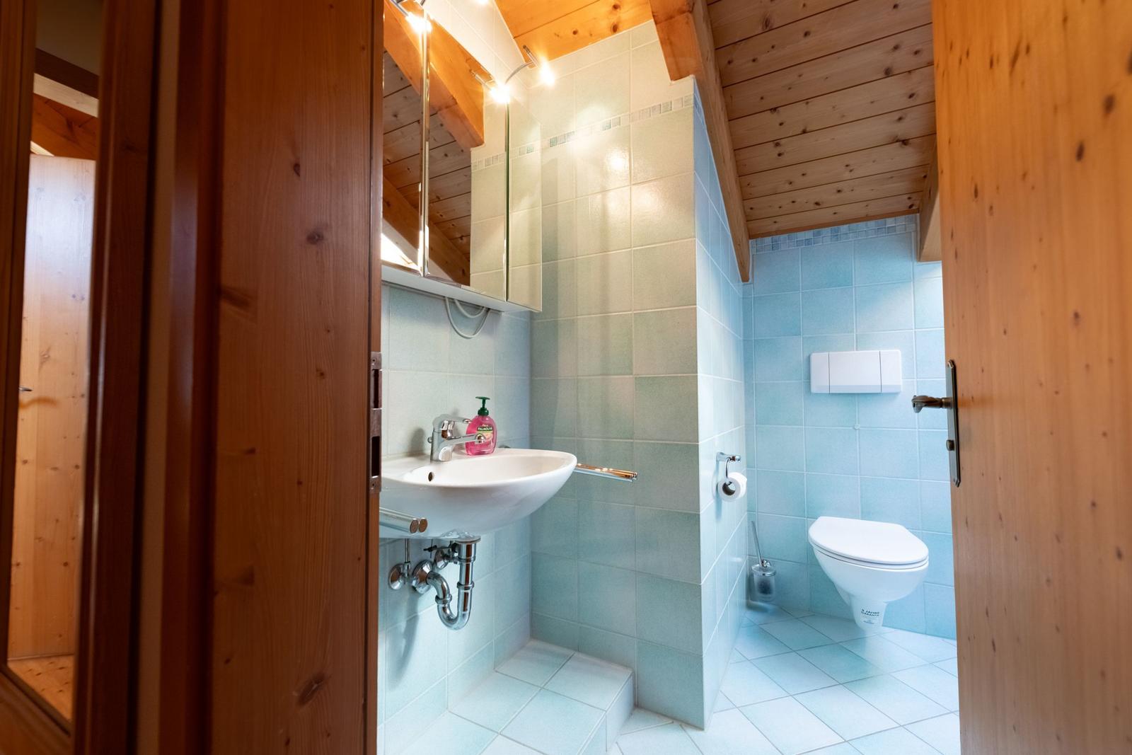 Badezimmer mit Waschbecken, Toilette und Dusche. Holzdecke und hellblaue Fliesen.