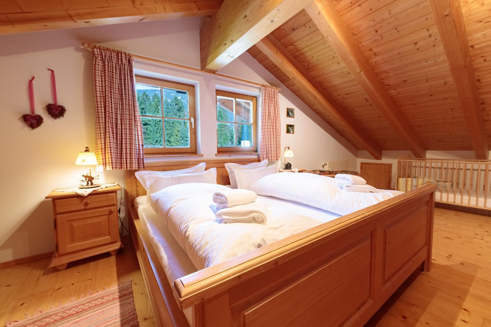 Schlafzimmer mit Holzbett, zwei Fenstern und Holzdecke. Zimmer ist mit Holzmöbeln eingerichtet.