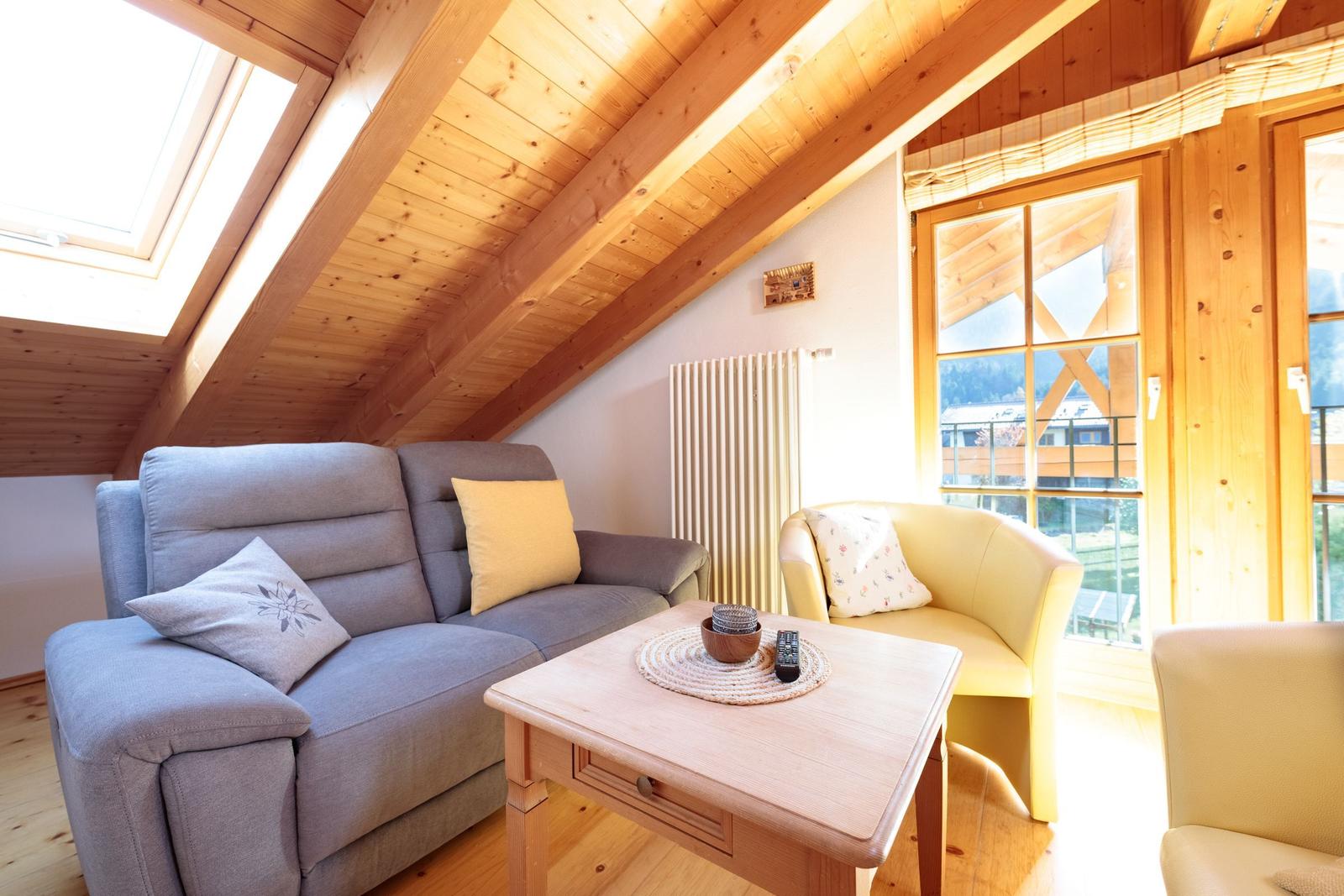 Wohnzimmer mit Sofa, Sitzgelegenheiten und Holzdecke. Fenster und Dachfenster bieten Blick auf die Umgebung.