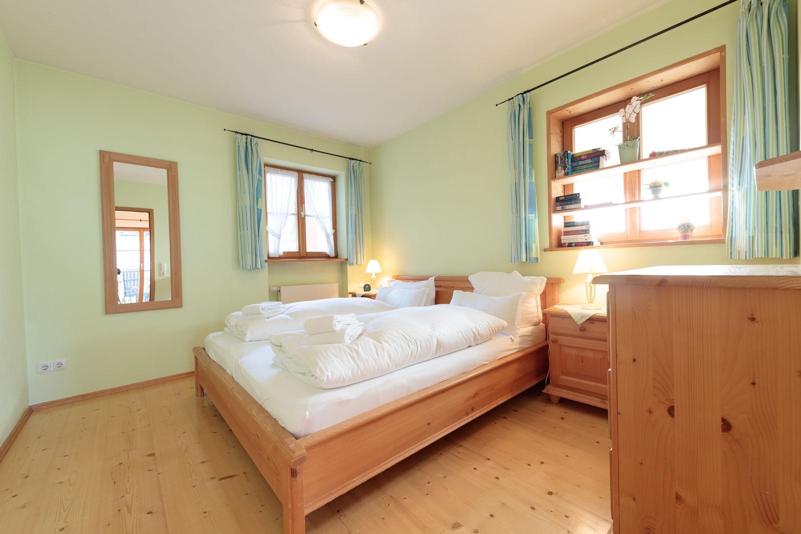Schlafzimmer mit Holzbett, Fenster und Schrank. Wandspiegel und Nachttisch mit Lampe.