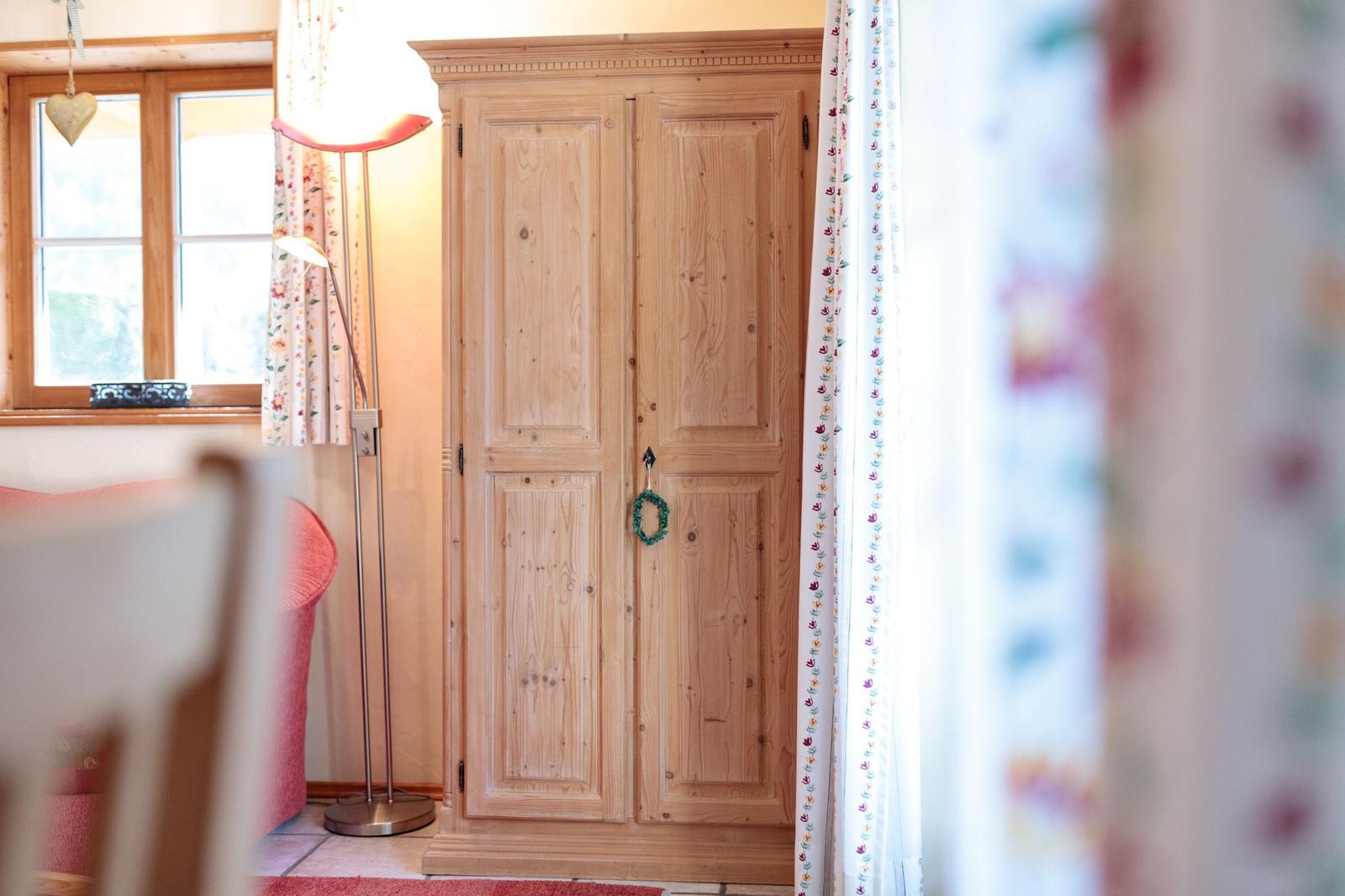 Holzschrank mit grüner Girlande, Fenster mit Vorhängen und Stehlampe im Raum.