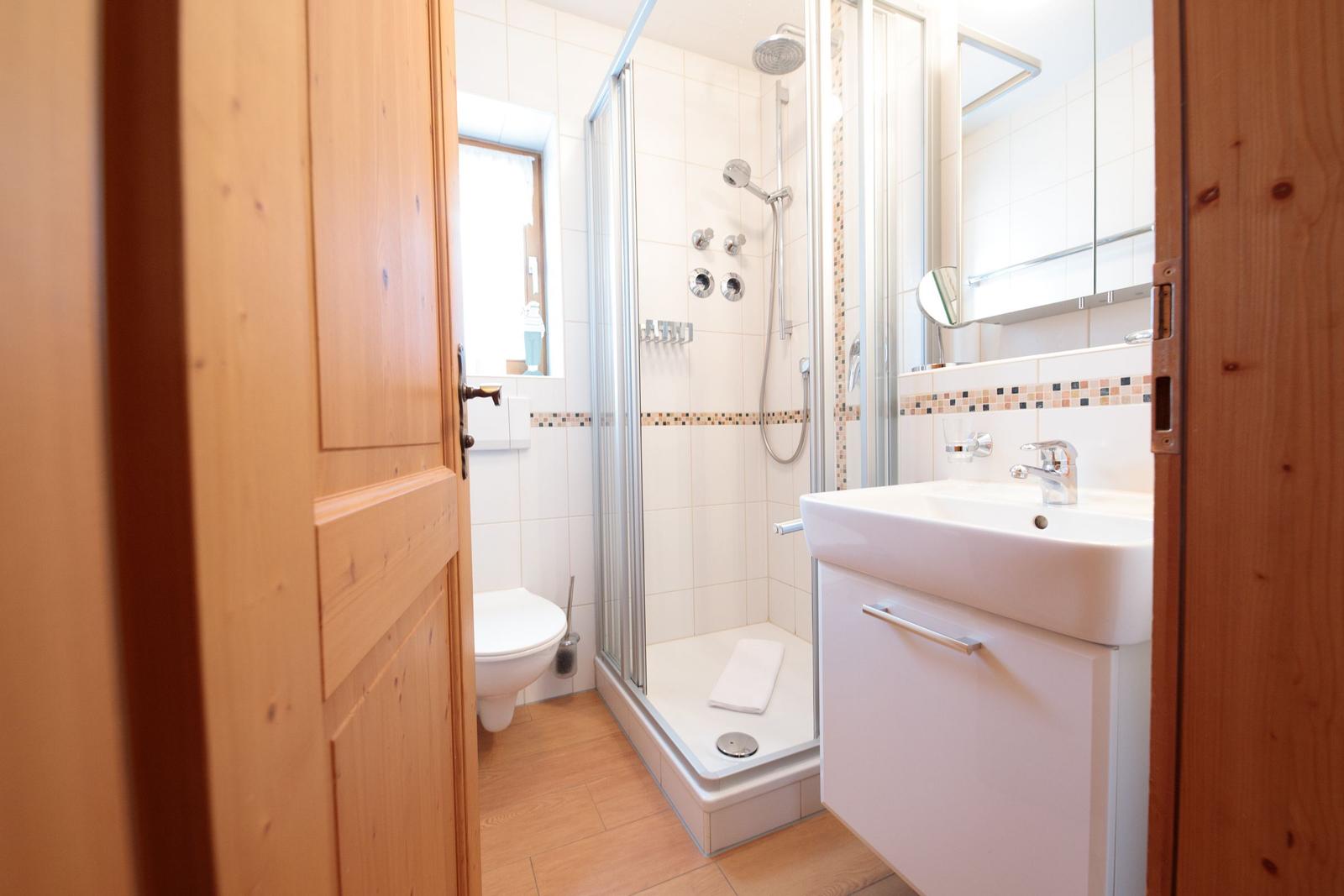 Badezimmer mit Dusche, Waschbecken und Toilette. Holztür und hell gefliest.
