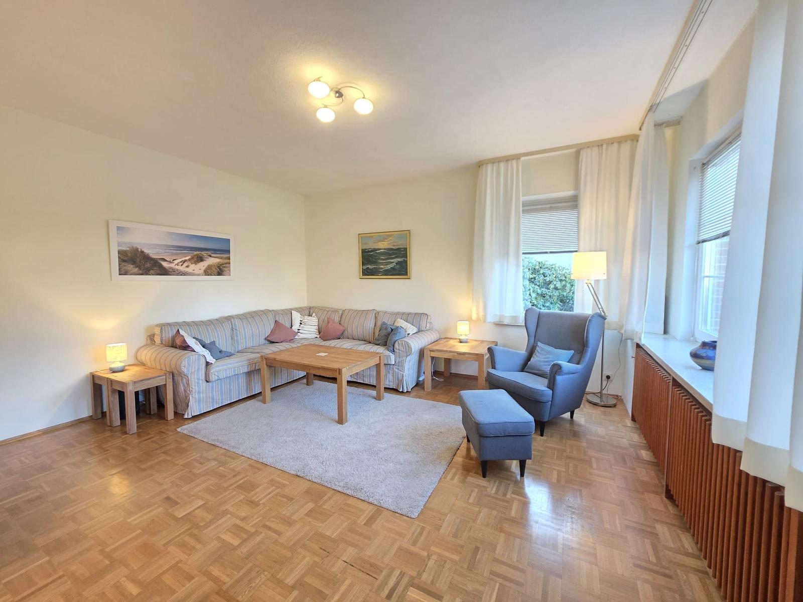 Wohnzimmer mit Sofa, Sessel, Tisch und Fenstern.