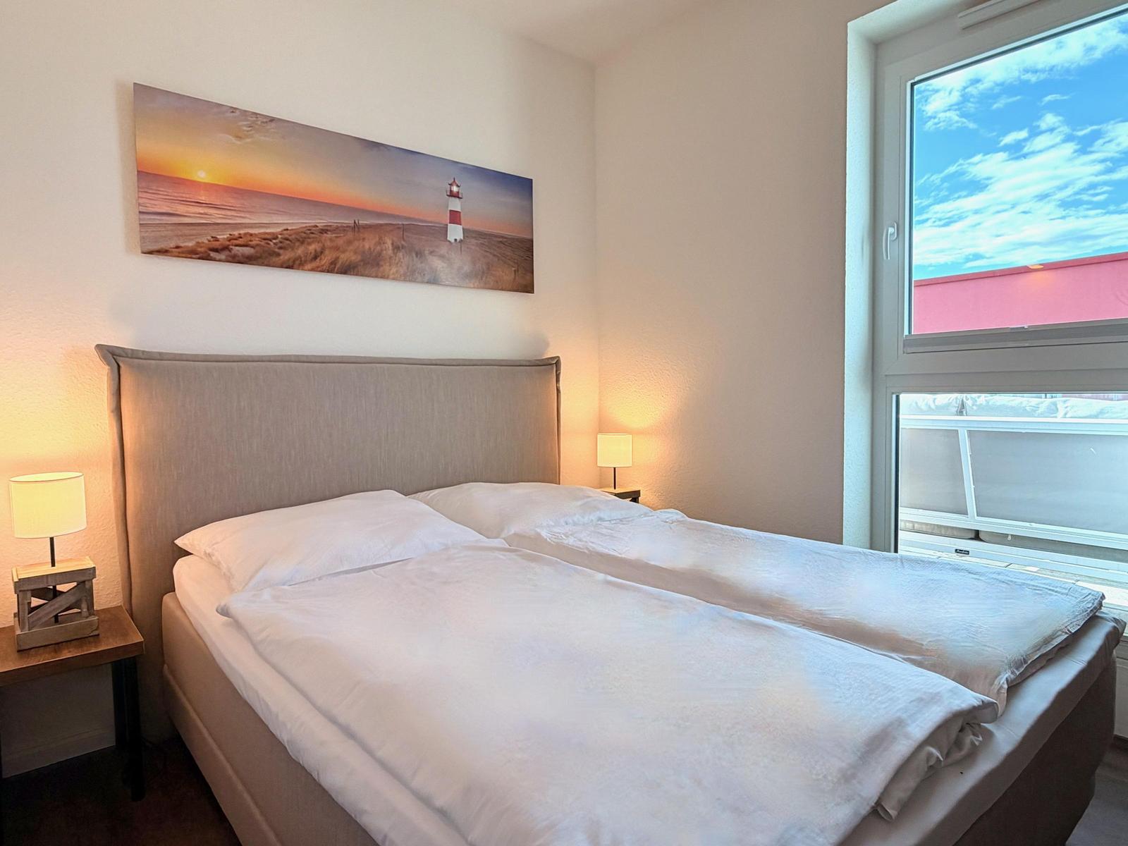 Schlafzimmer mit Bett, Nachttischen und Wandbild. Fenster zeigt Außenbereich.