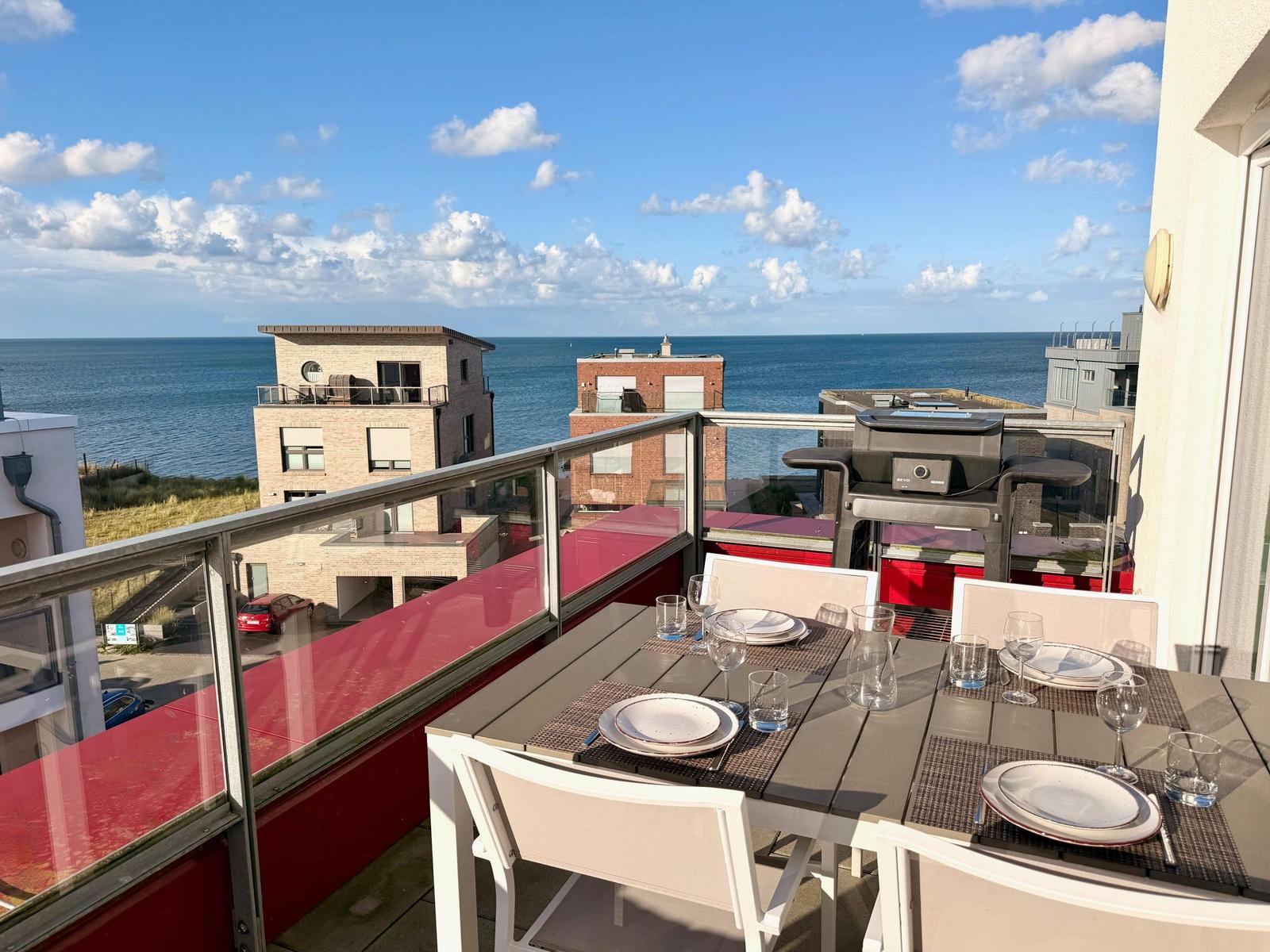 Terrasse mit Tisch, Stühlen und Grill. Blick auf Meer und Gebäude.