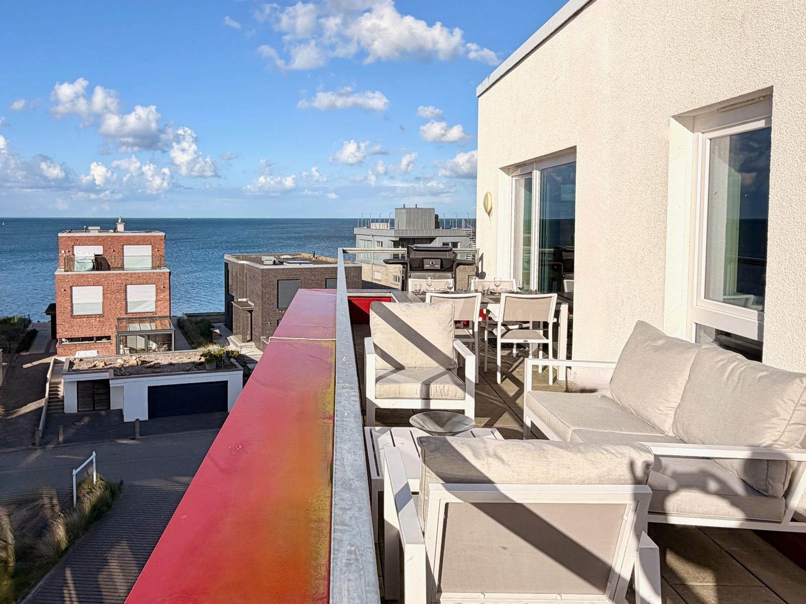 Terrasse mit Sitzgelegenheiten und Meerblick. Rote Balustrade, moderne Möbel.