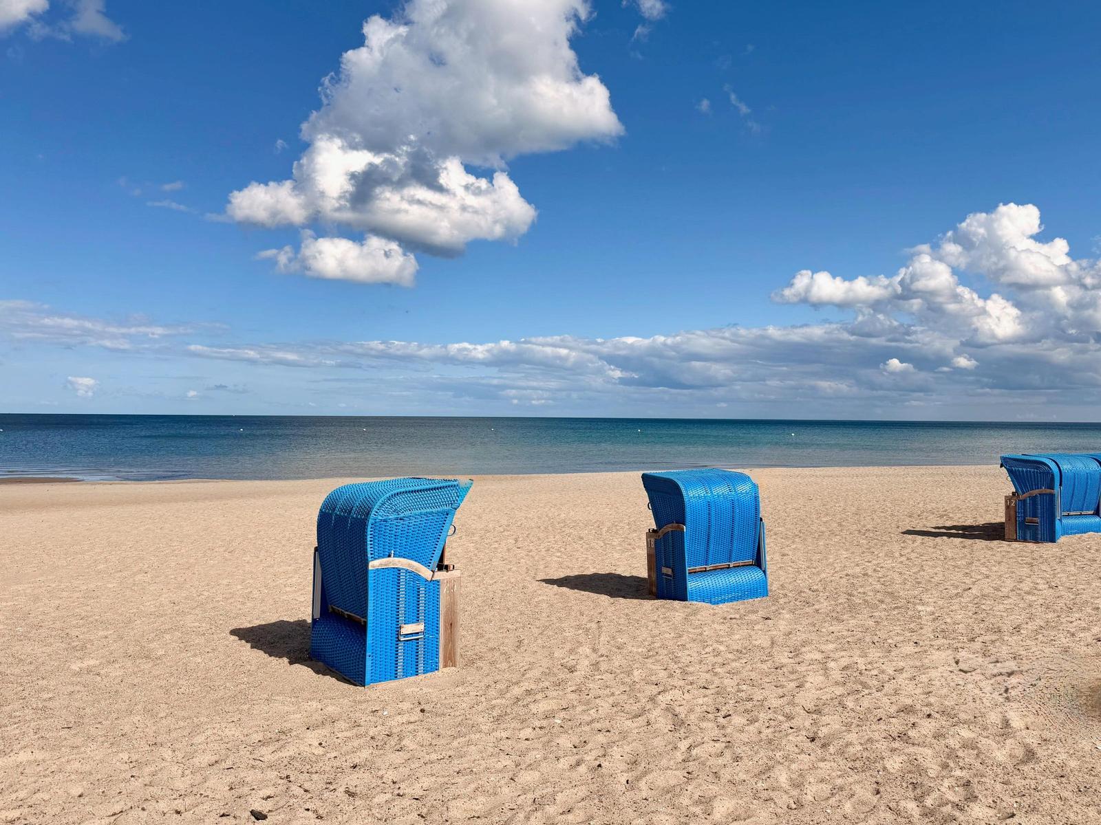 Drei blaue Strandkörbe stehen auf sandigem Strand vor dem Meer unter blauem Himmel mit weißen Wolken.