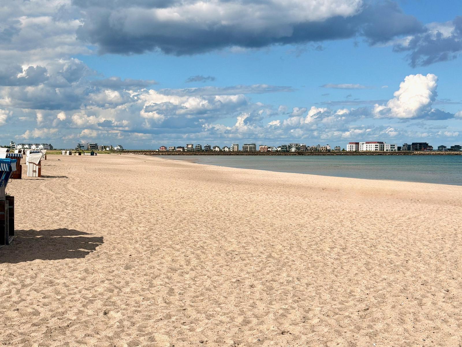 Strand mit Sand, Strandkörben und Häusern am Horizont unter bewölktem Himmel.