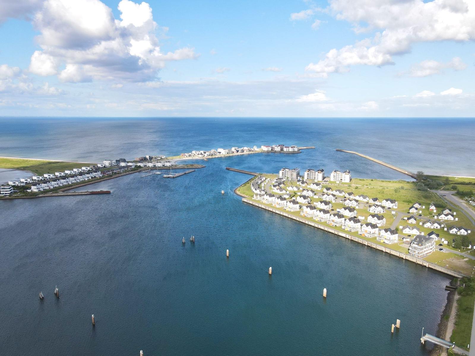 Aerial view of coastal town with marina, houses, and sea under partly cloudy sky.