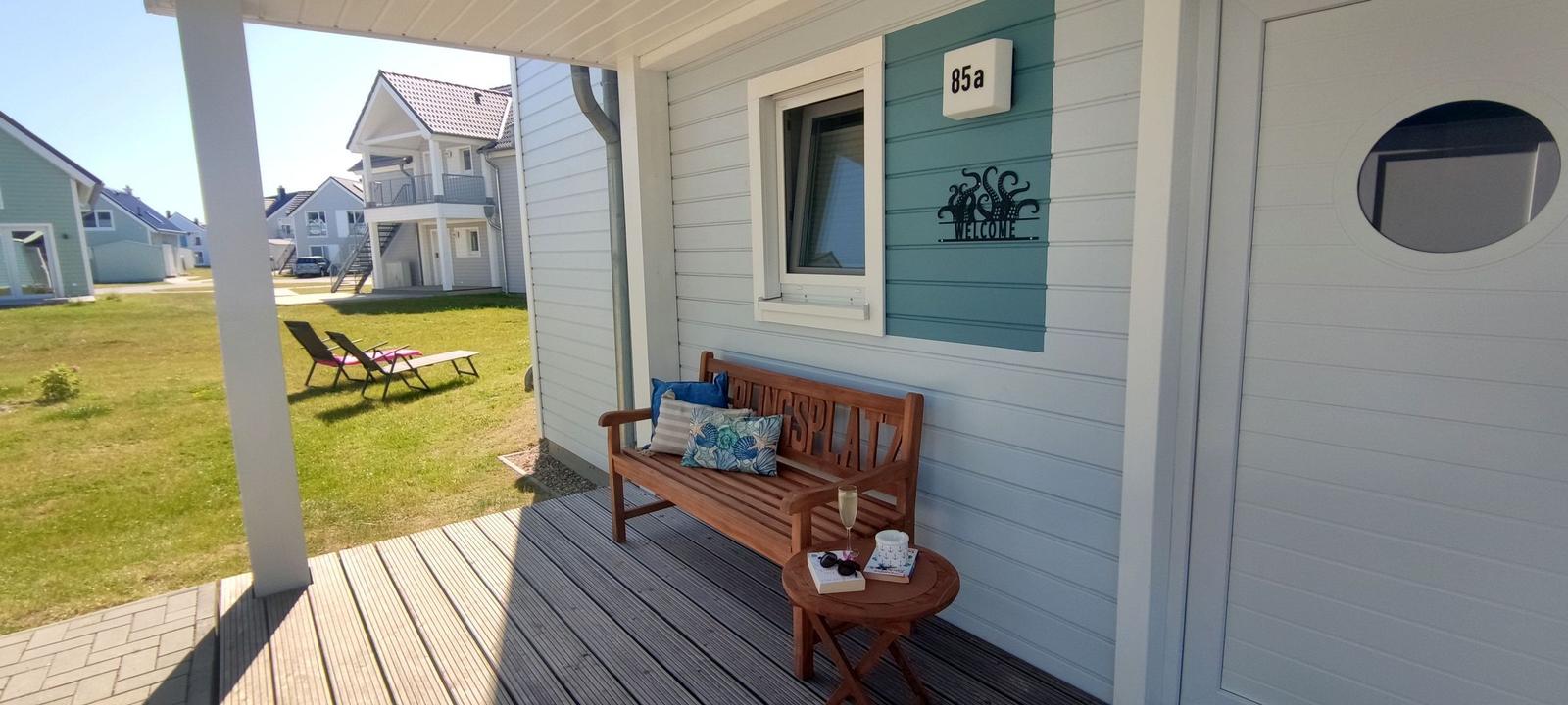 Deck with wooden bench, table, and drinks in front of a house with blue siding and round door window.
