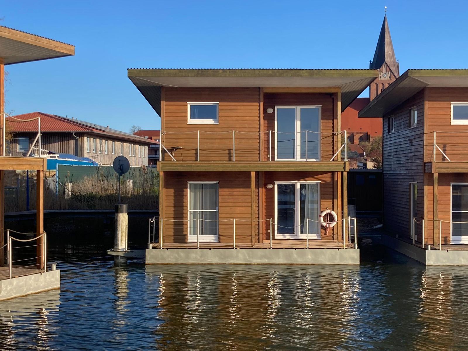 Holzhaus auf Wasser mit Balkon und Fenstern. Im Hintergrund Kirchturm.