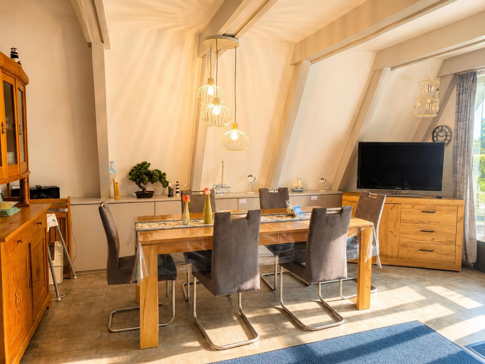 Esszimmer mit Holztisch, Stühlen und Deckenleuchten. Fernseher und Fenster sichtbar.