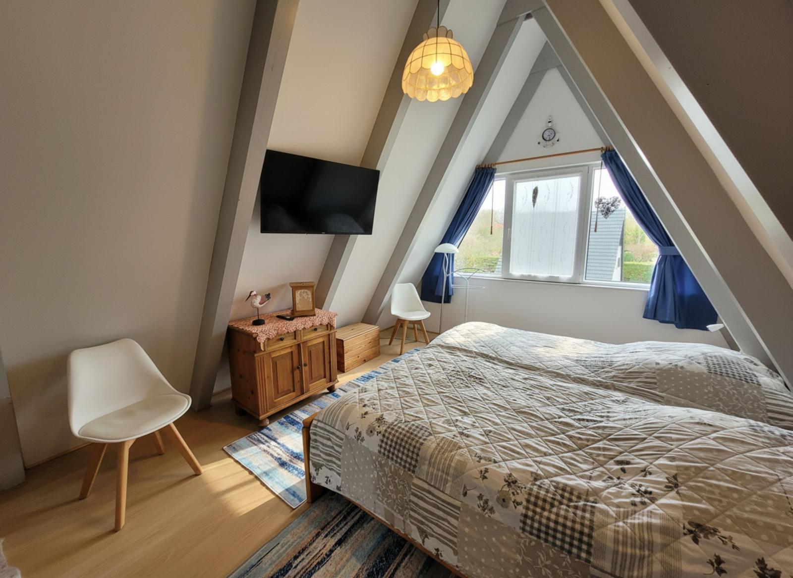 Schlafzimmer mit Dachschrägen, Bett, TV und Fenster mit blauen Vorhängen.