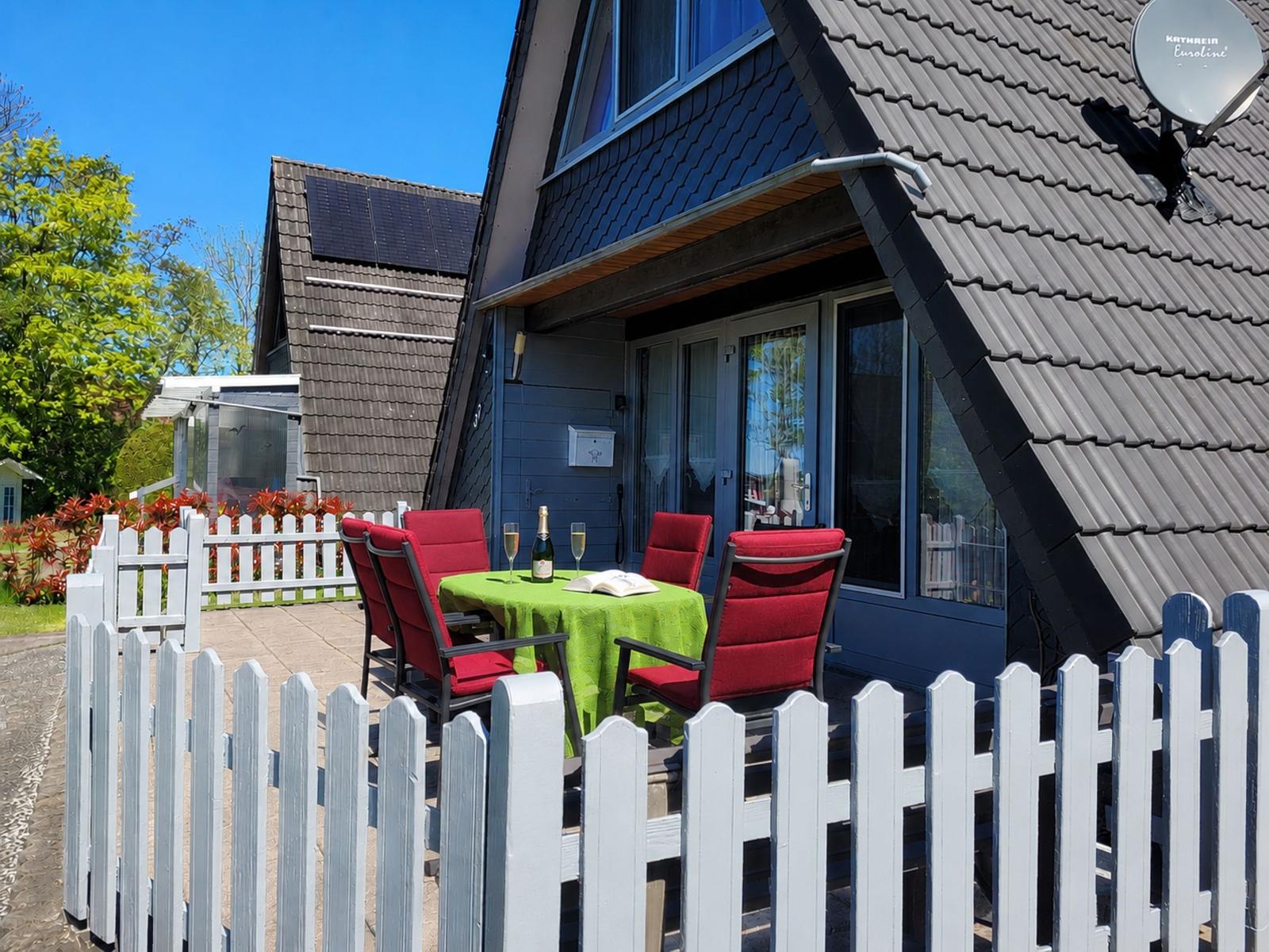 Terrasse mit Tisch, Stühlen und Wein. Haus mit grauem Dach und weißem Zaun.