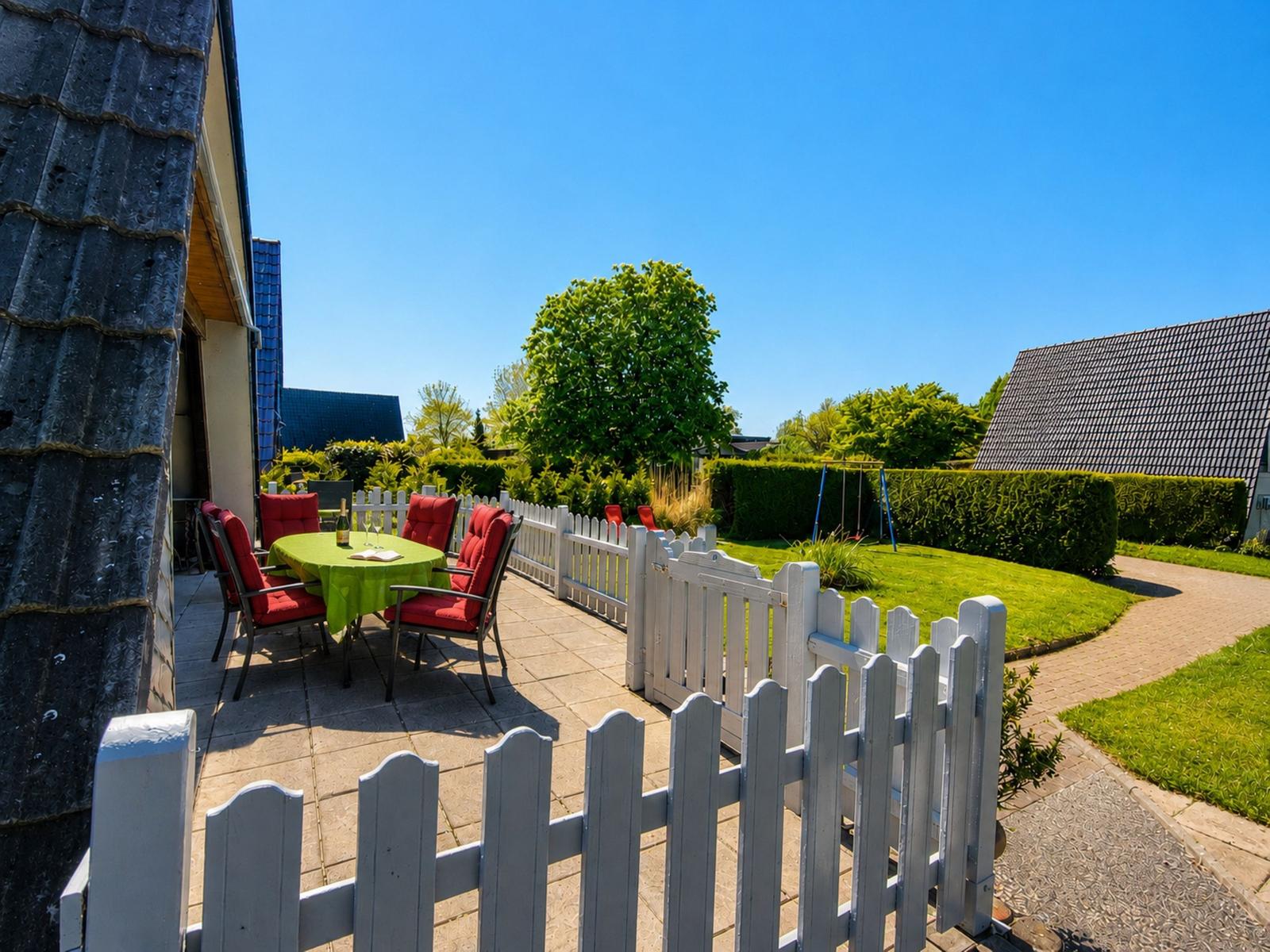 Terrasse mit Tisch und Stühlen, weißer Zaun, Garten und Spielplatz im Hintergrund.