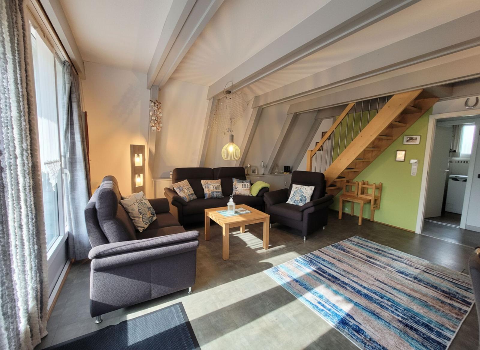Wohnzimmer mit Sofa, Sessel und Holztisch. Treppenhaus und Fenster sichtbar.