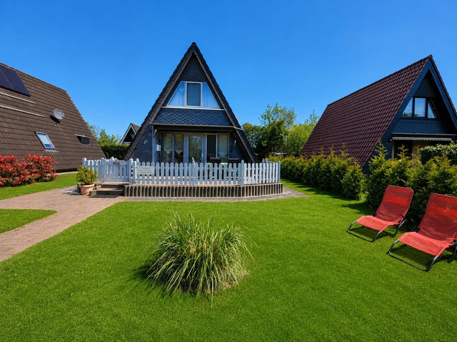 A-frame-Haus mit Garten, weiße Umzäunung und zwei roten Liegestühlen.