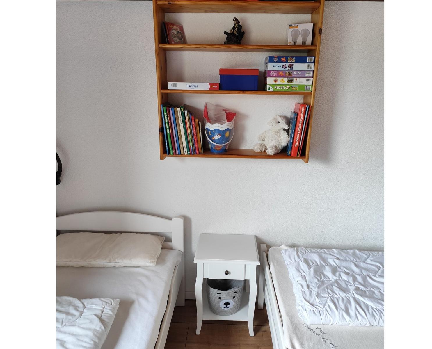Two beds with a nightstand and wall shelf with books and toys.