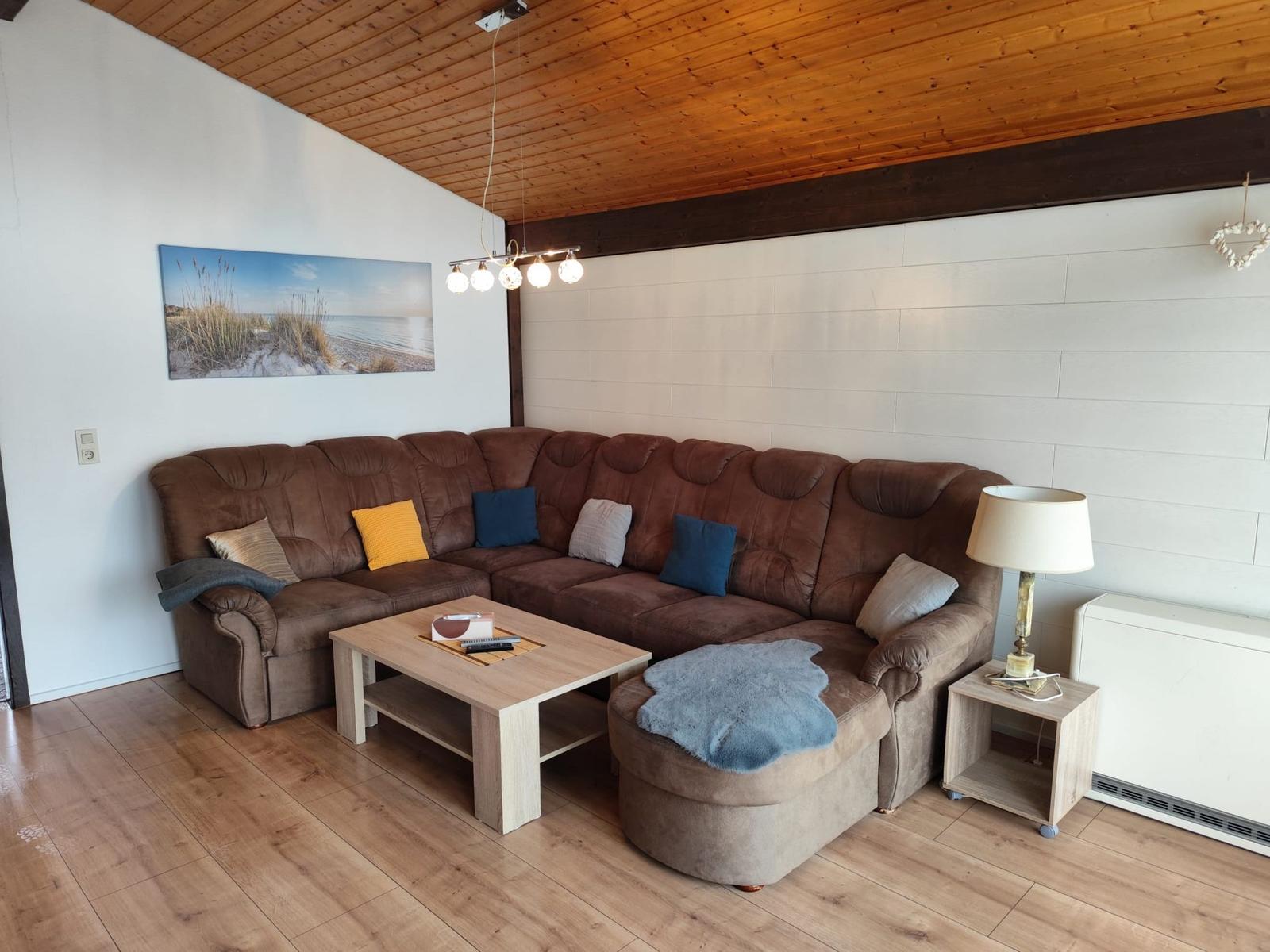 A living room with a brown sofa, wooden floor, and wooden ceiling. There is a coffee table and a table lamp.