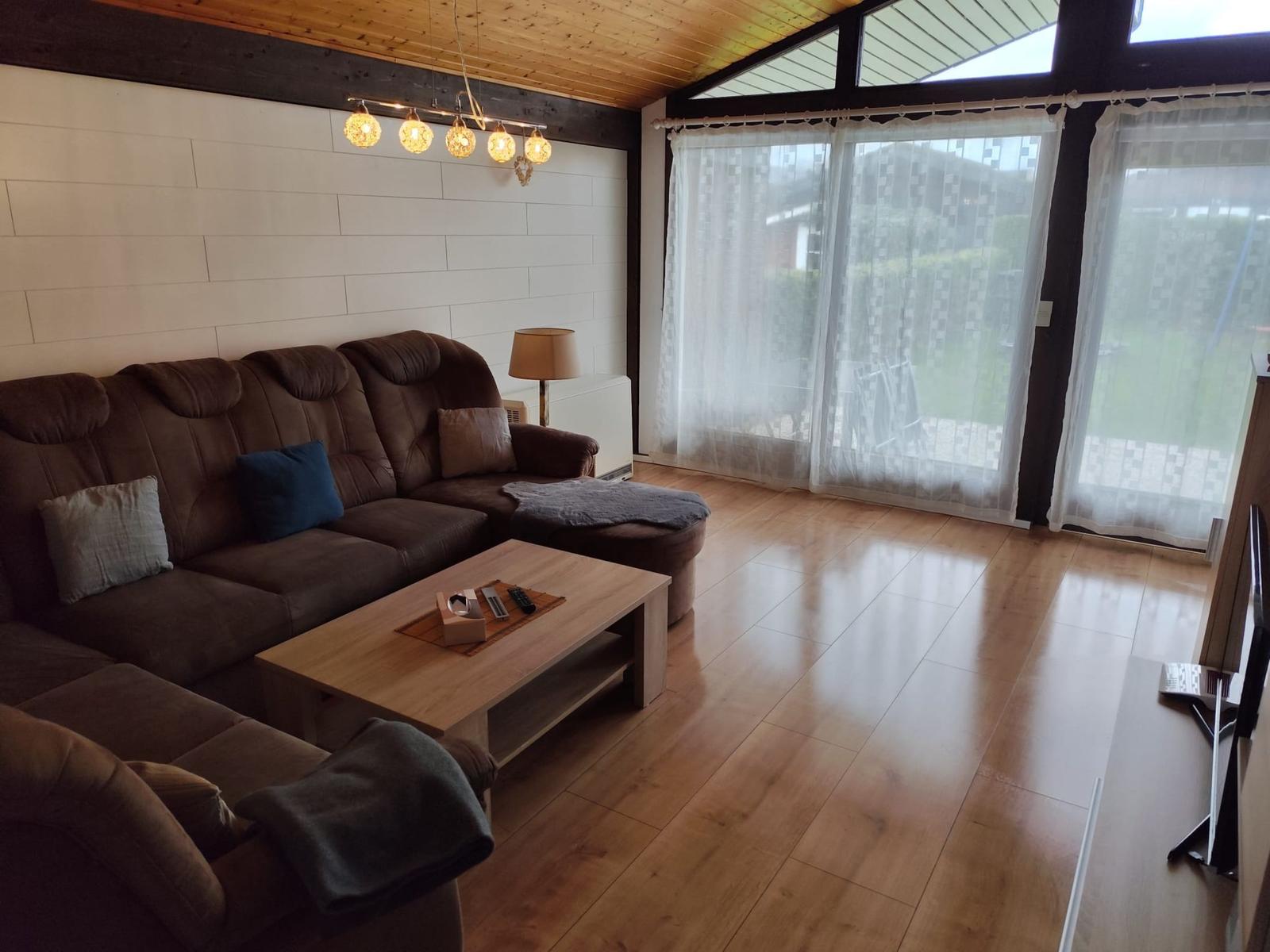 A living room with a brown sofa, wooden floor, and large windows with white curtains.