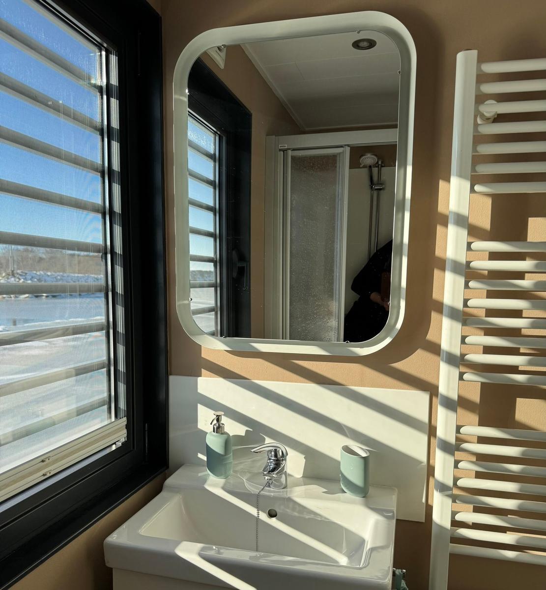 Badezimmer mit Waschbecken, Spiegel und Dusche. Fenster mit Jalousie und Heizkörper.