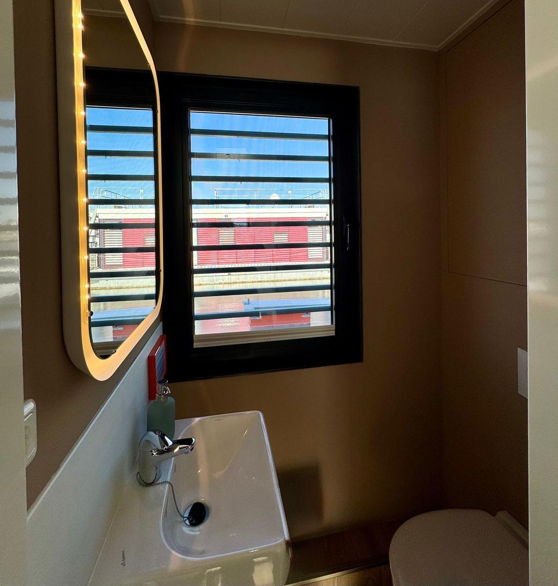 Badezimmer mit Waschbecken, Spiegel und Fenster mit Jalousie.