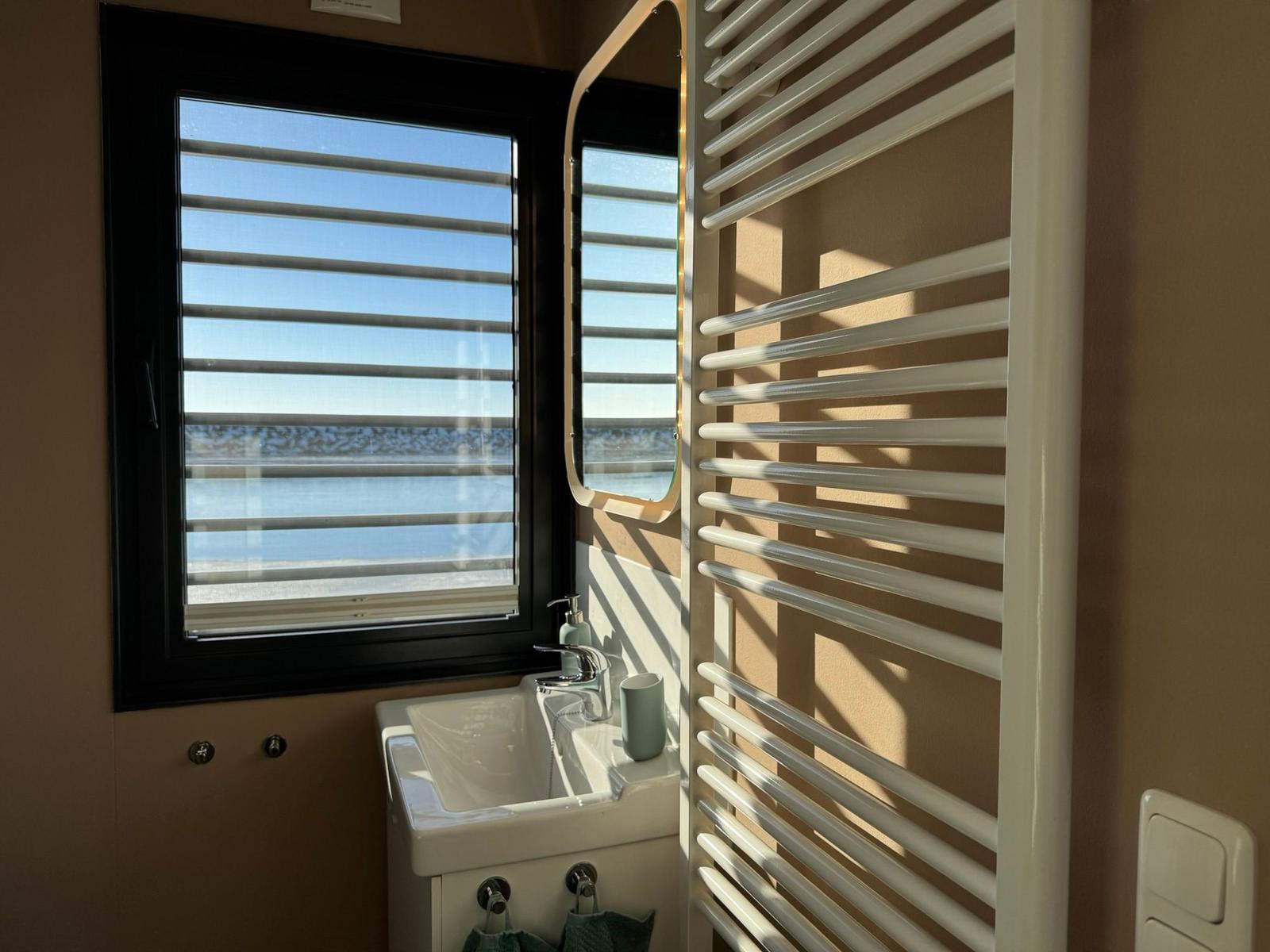 Badezimmer mit Waschbecken, Spiegel und Heizkörper. Fenster mit Jalousie zeigt Blick auf Wasser.