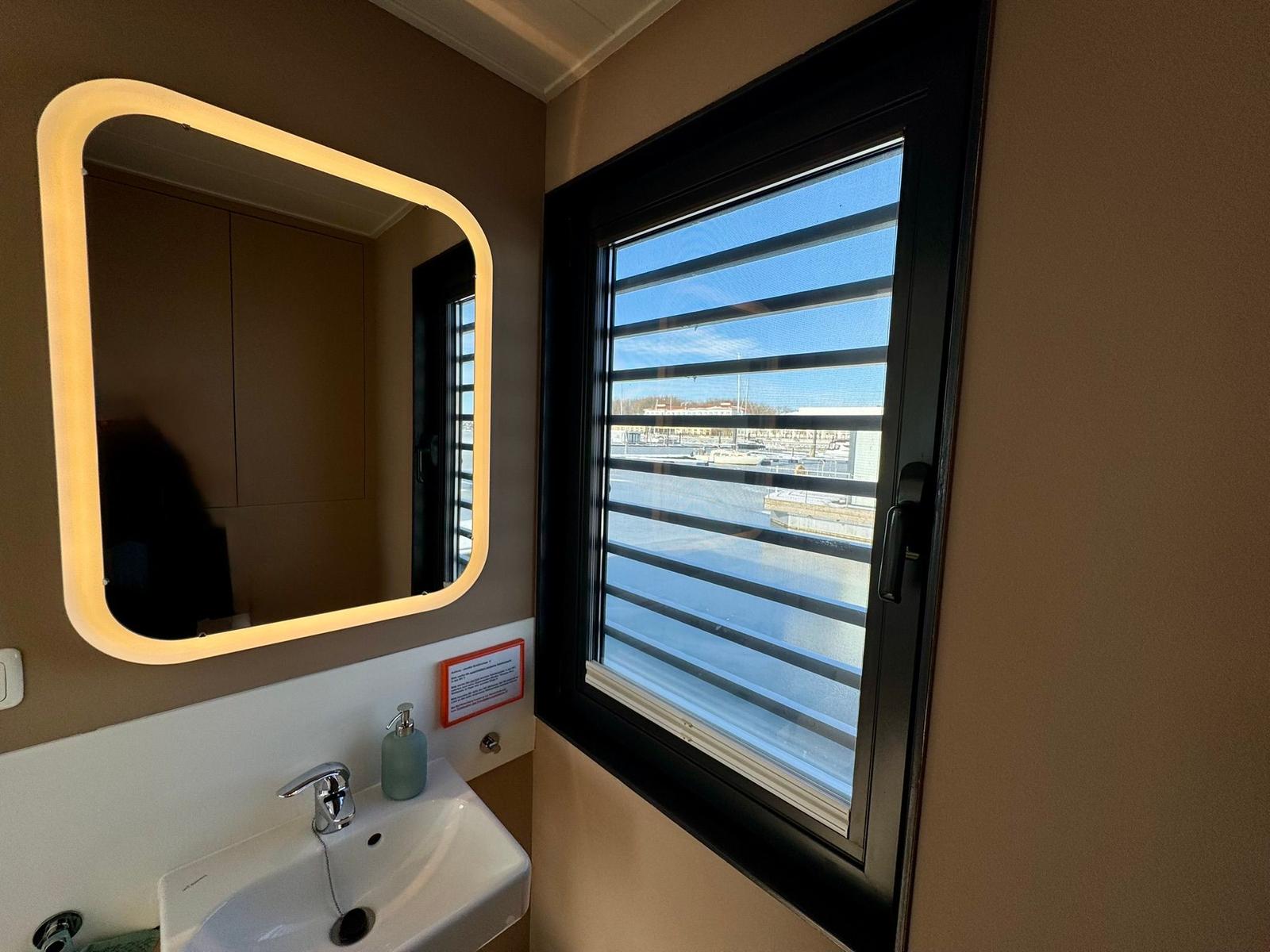 Badezimmer mit Waschbecken, beleuchtetem Spiegel und Fenster mit Blick auf Hafen.