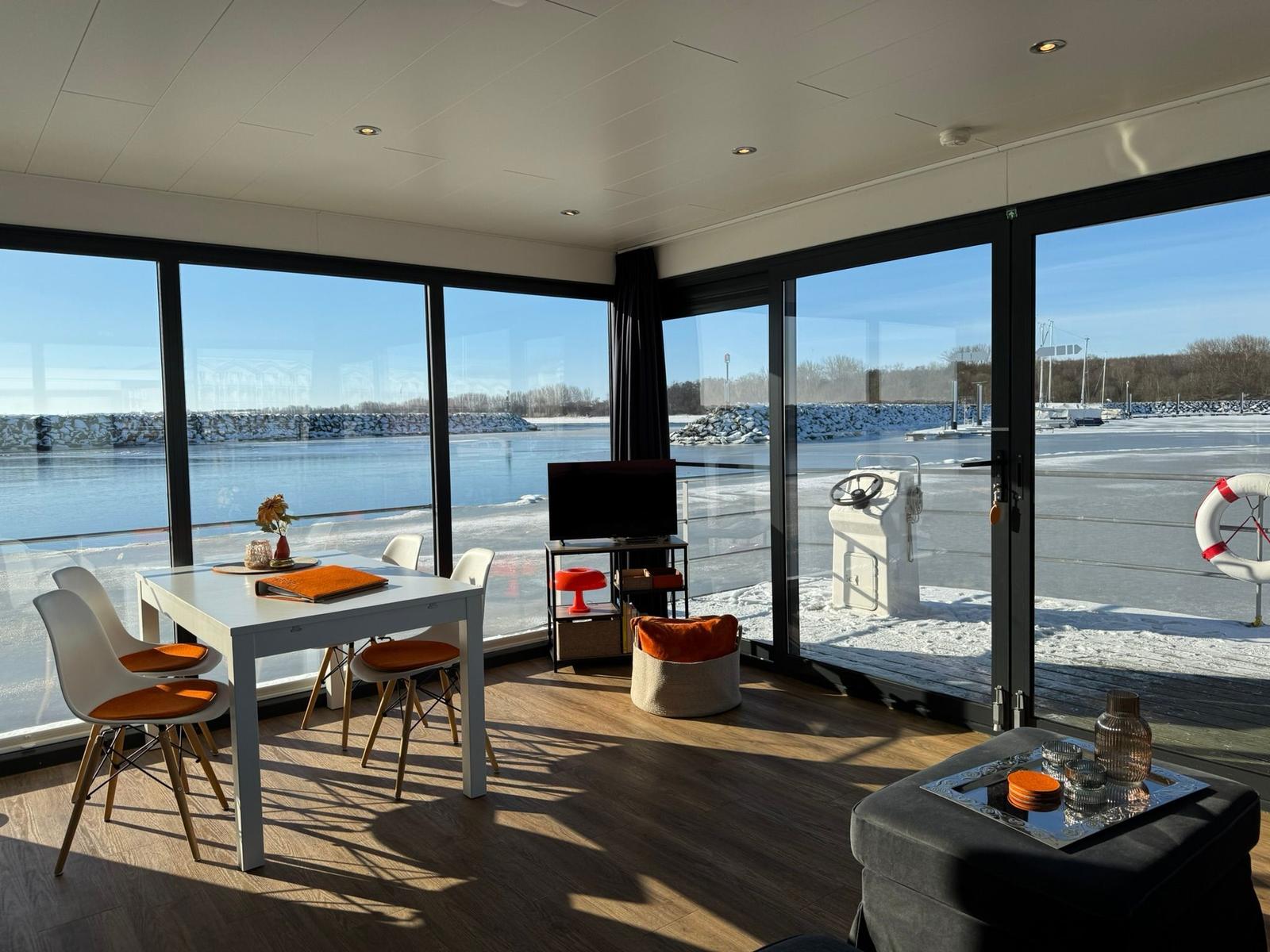 Ein Raum mit großem Fensterblick auf gefrorenen Hafen. Esstisch, Sessel und Fernseher sind vorhanden.