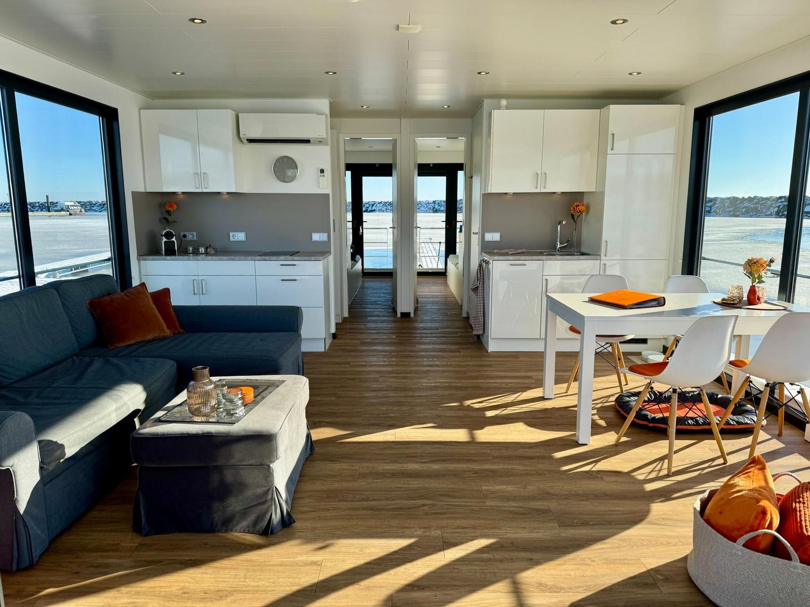 Wohnung mit Küchenbereich, Esstisch und Sofa. Große Fenster mit Blick auf Wasser.